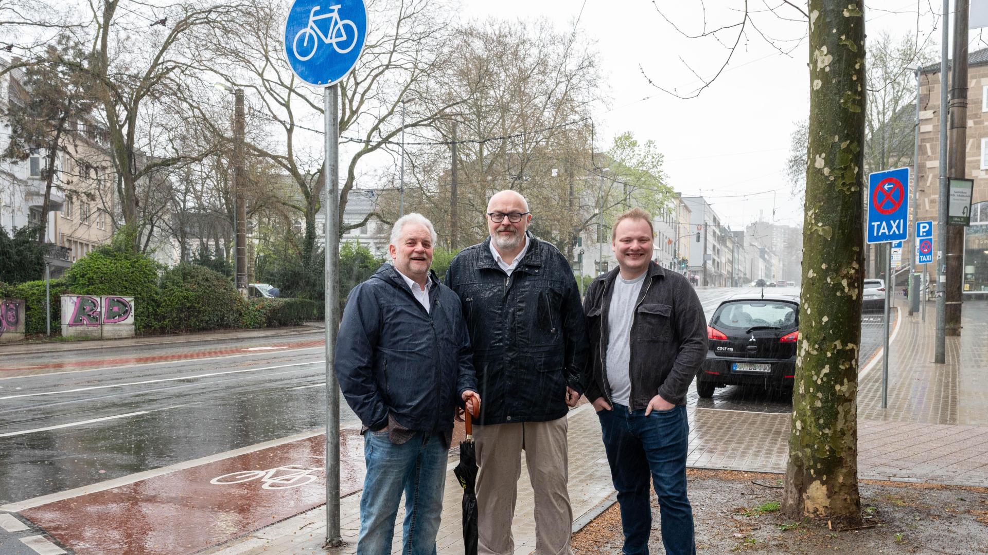 Drei Männer stehen auf einem Bürgersteig neben dem fertigen Radweg an der Kaiserstraße mit Fahrradpiktogramm und blauem Fahrradverkehrsschild bei regnerischem Wetter.