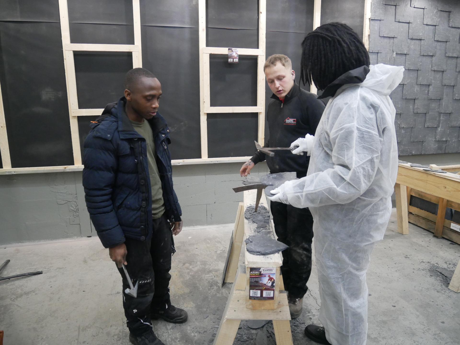 Jugendliche bekommen in einer Lehrwerkstatt gezeigt, wie sie auf einem Holzbock mit entsprechendem Werkzeug aus einer Schieferplatte ein Herz schlagen.