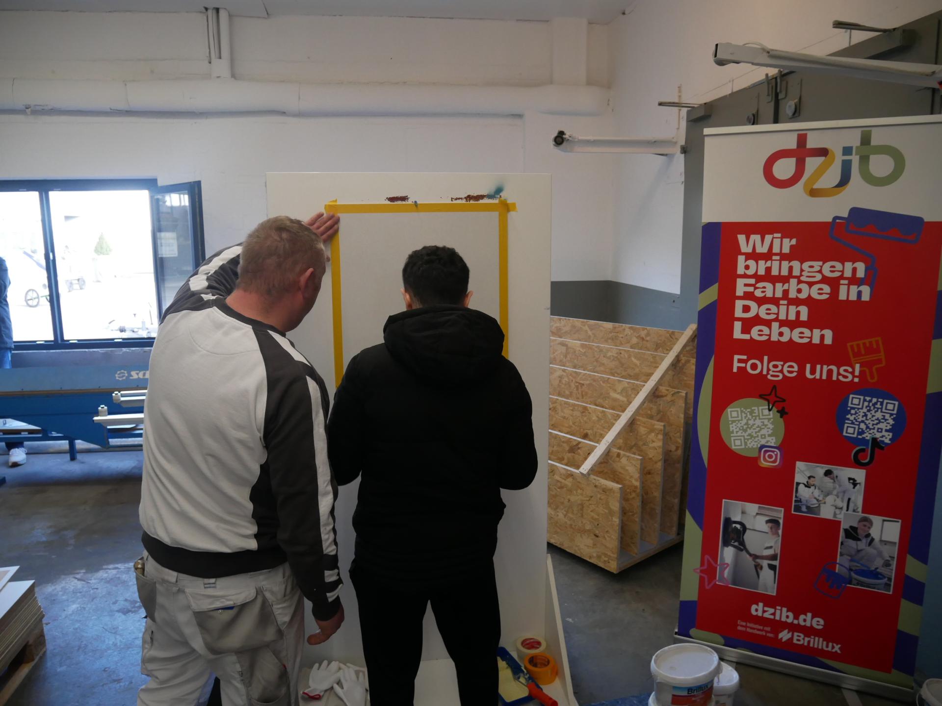 Ein Mitarbeiter eines Malerbetriebs zeigt einem Jugendlichen an einer abgeklebten Stellwand, wie er diese richtig streicht.