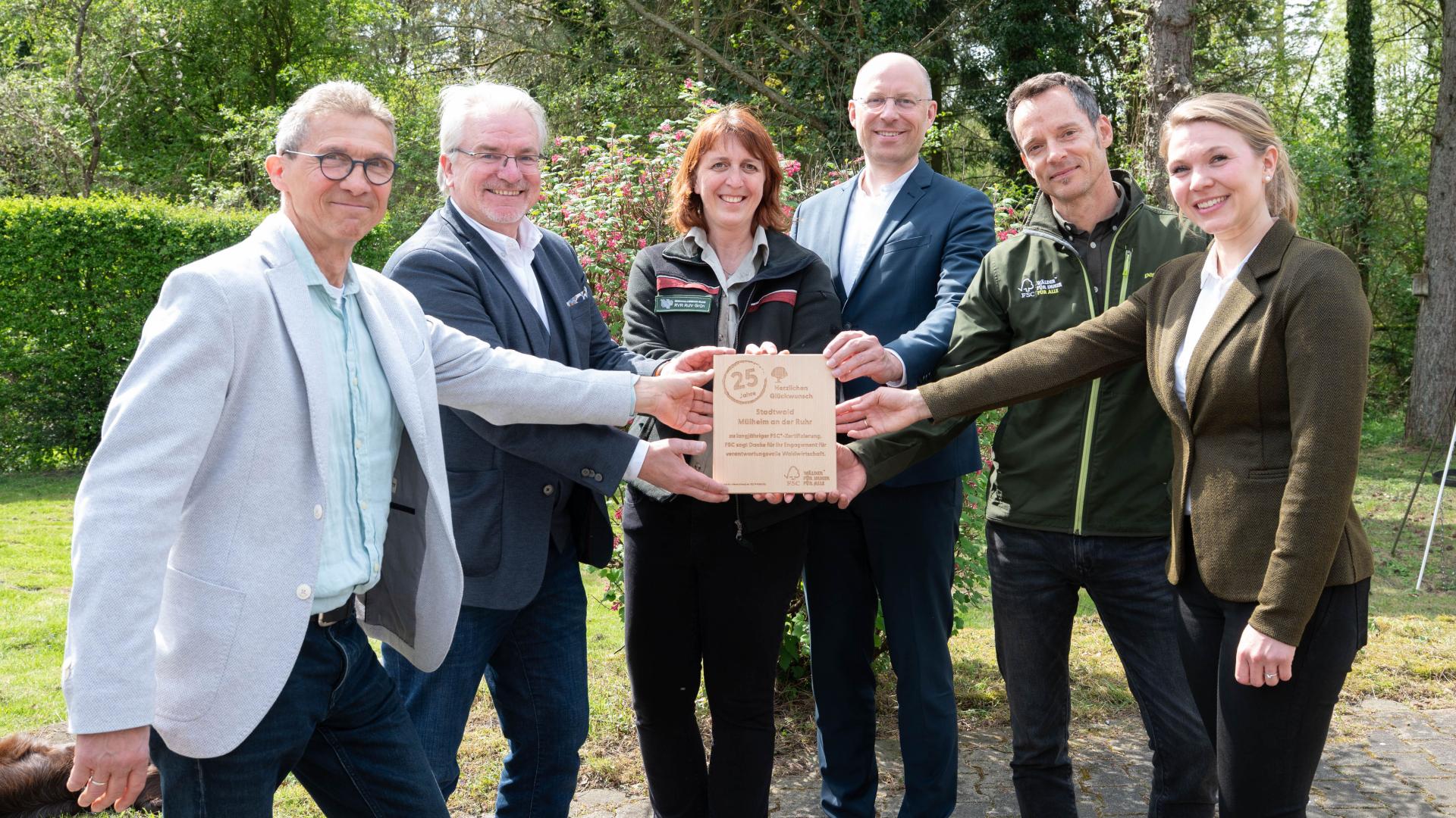 Sechs Personen stehen im Wald und halten gemeinsam eine Holztafel mit der FSC Jubiläumsgravur