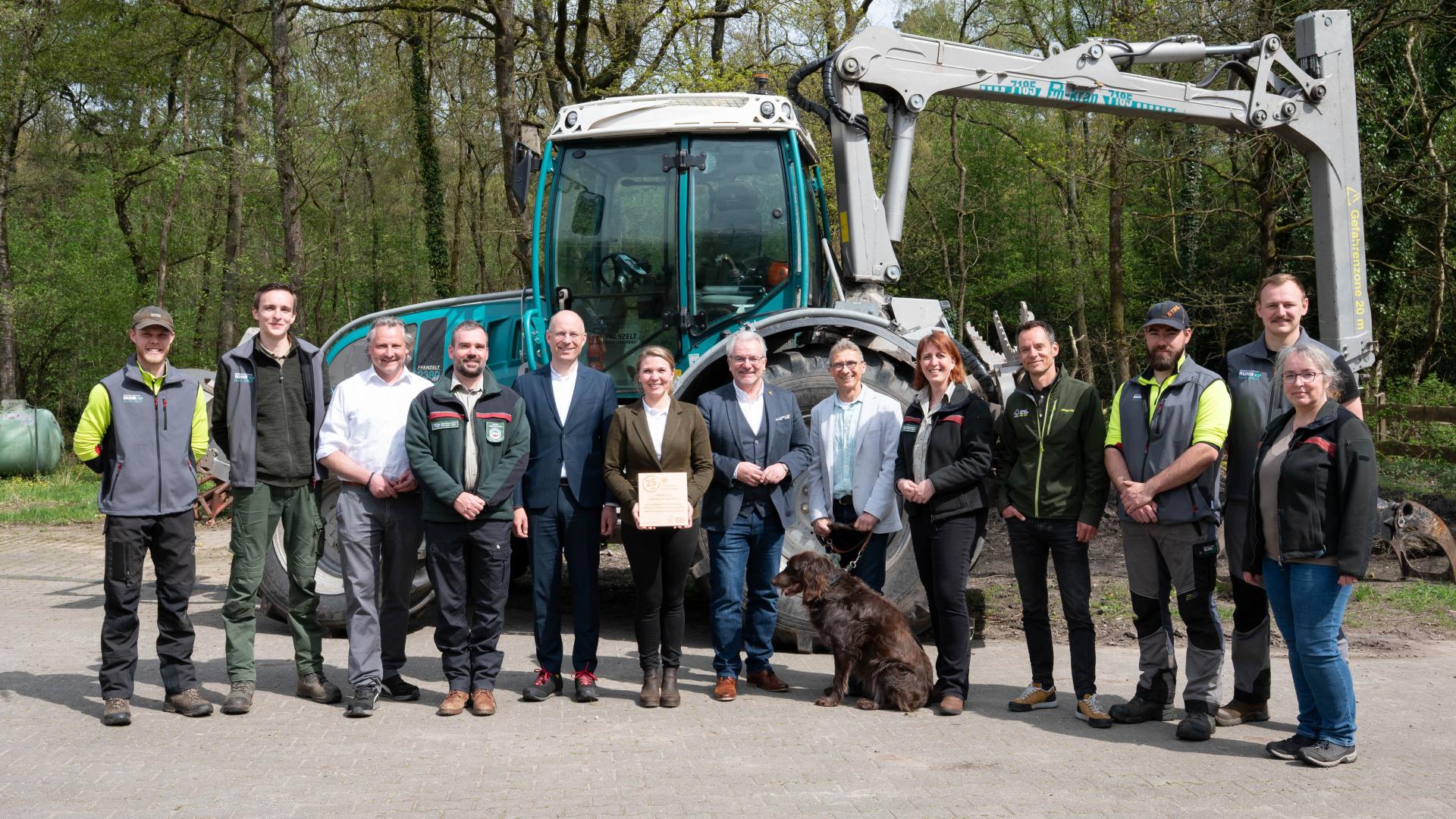 Gruppe von Menschen steht vor einem Traktor mit Frontlader im Mülheimer Wald, eine Person hält die FSC Urkunde, ein Hund sitzt vorne in der Mitte