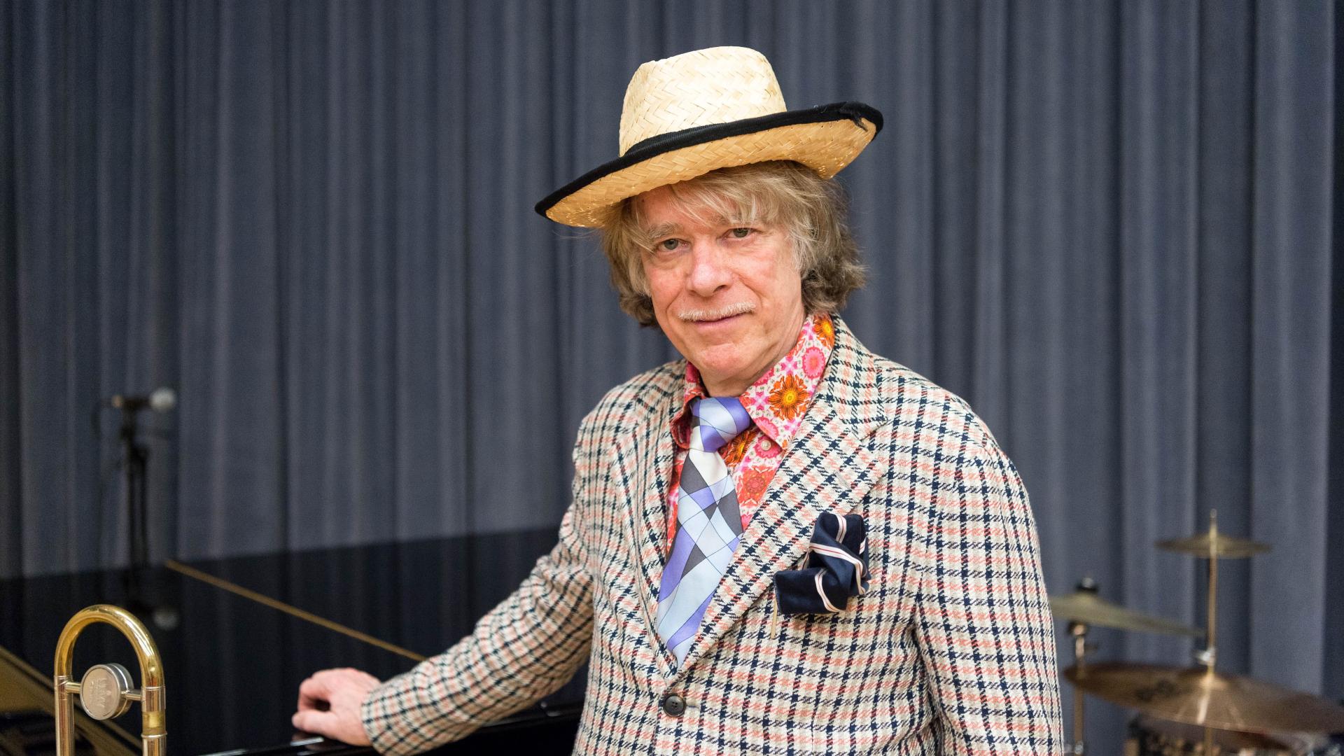 Helge Schneider mit Strohhut, kariertem Sakko, gemustertem Hemd und Krawatte steht vor einem Schlagzeug in der Musikschule Mülheim an  der Ruhr