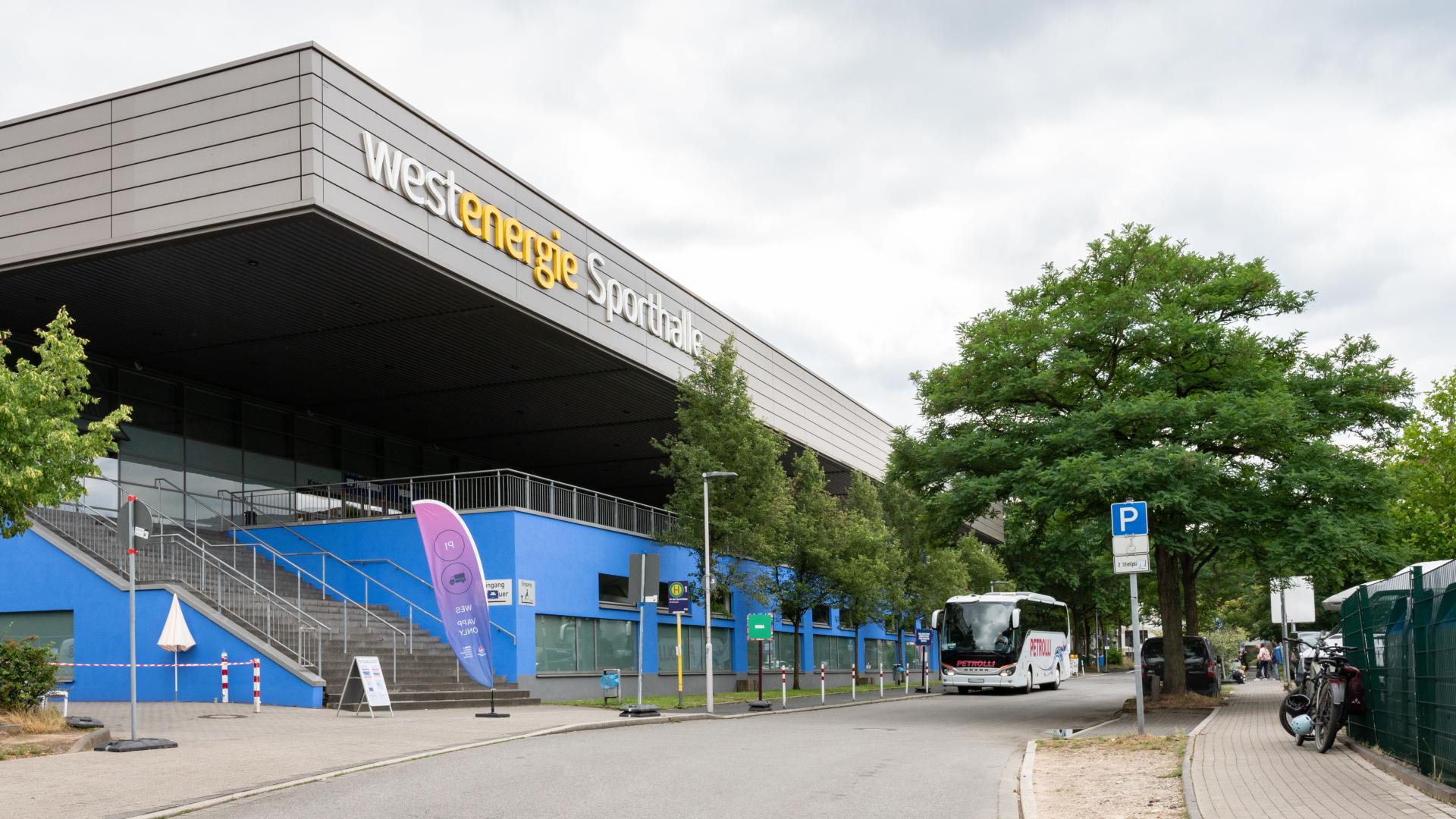 Außenansicht der Westenergie-Sporthalle mit blauem Sockel, Bäumen und einem Bus auf der Straße davor