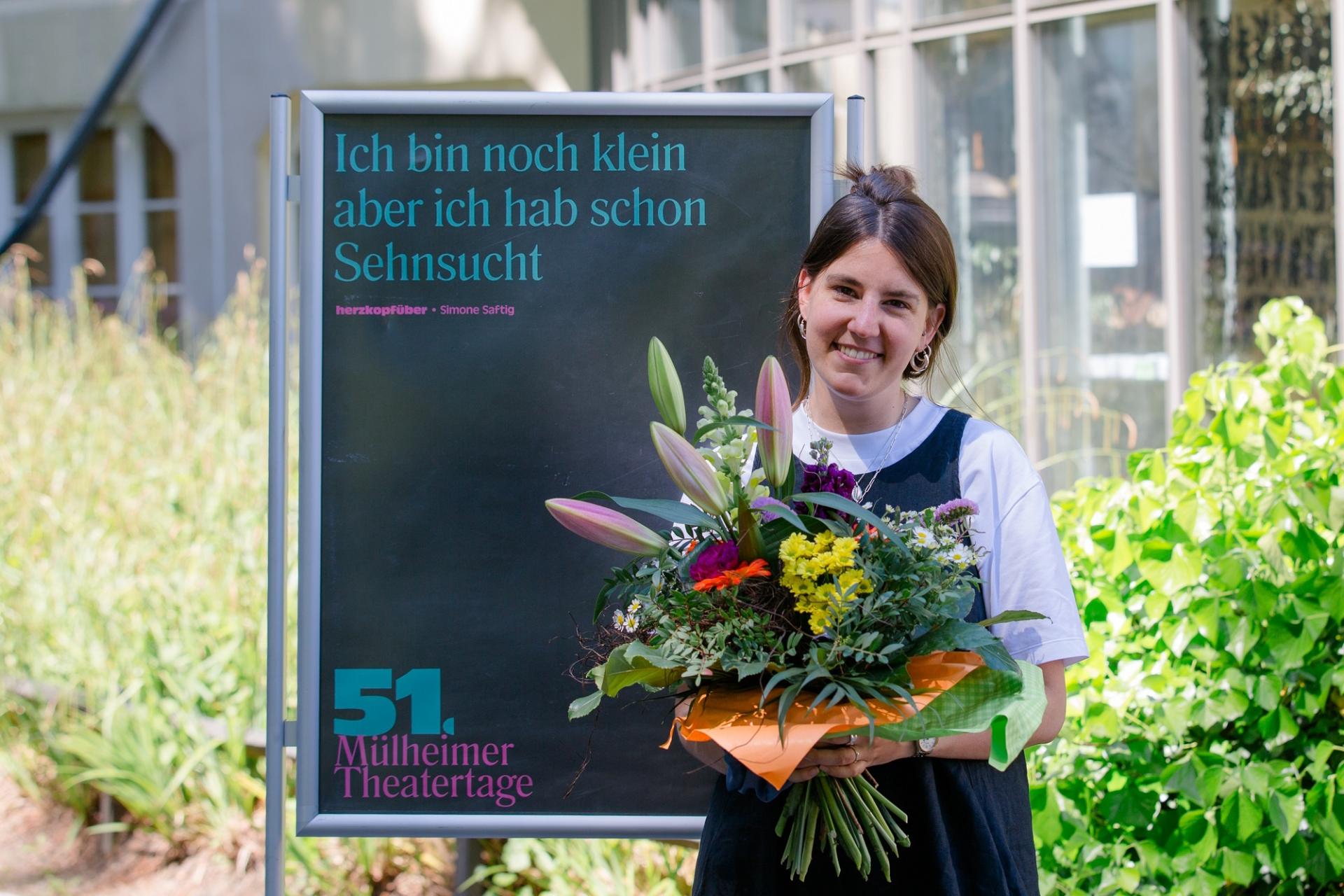 Die Autorin Simone Saftig steht mit einem Blumenstrauß vor einem Plakataufsteller.