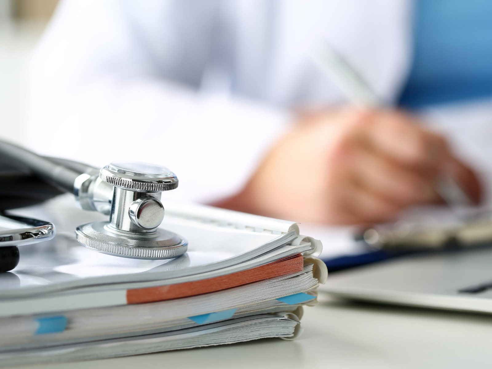 Der Bildausschnitt zeigt ein Stethoskop auf einem Stapel Unterlagen und im Hintergrund die Hand eines Arztes beim Handeintrag in einer Patientenakte.