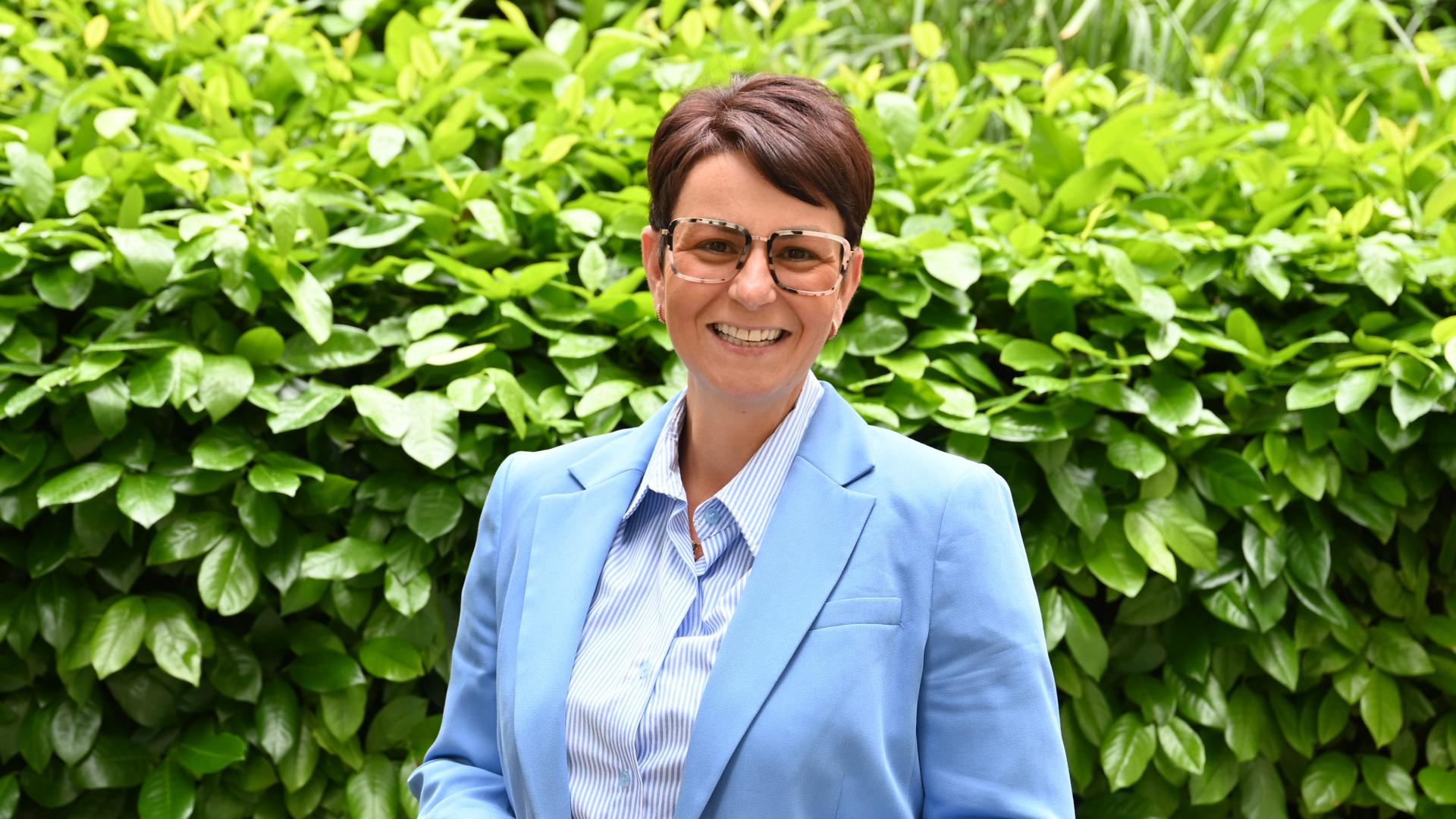 Person mit kurzen braunen Haaren trägt ein himmelblaues Jackett und ein weißes Oberteil, steht vor einer grünen Hecke