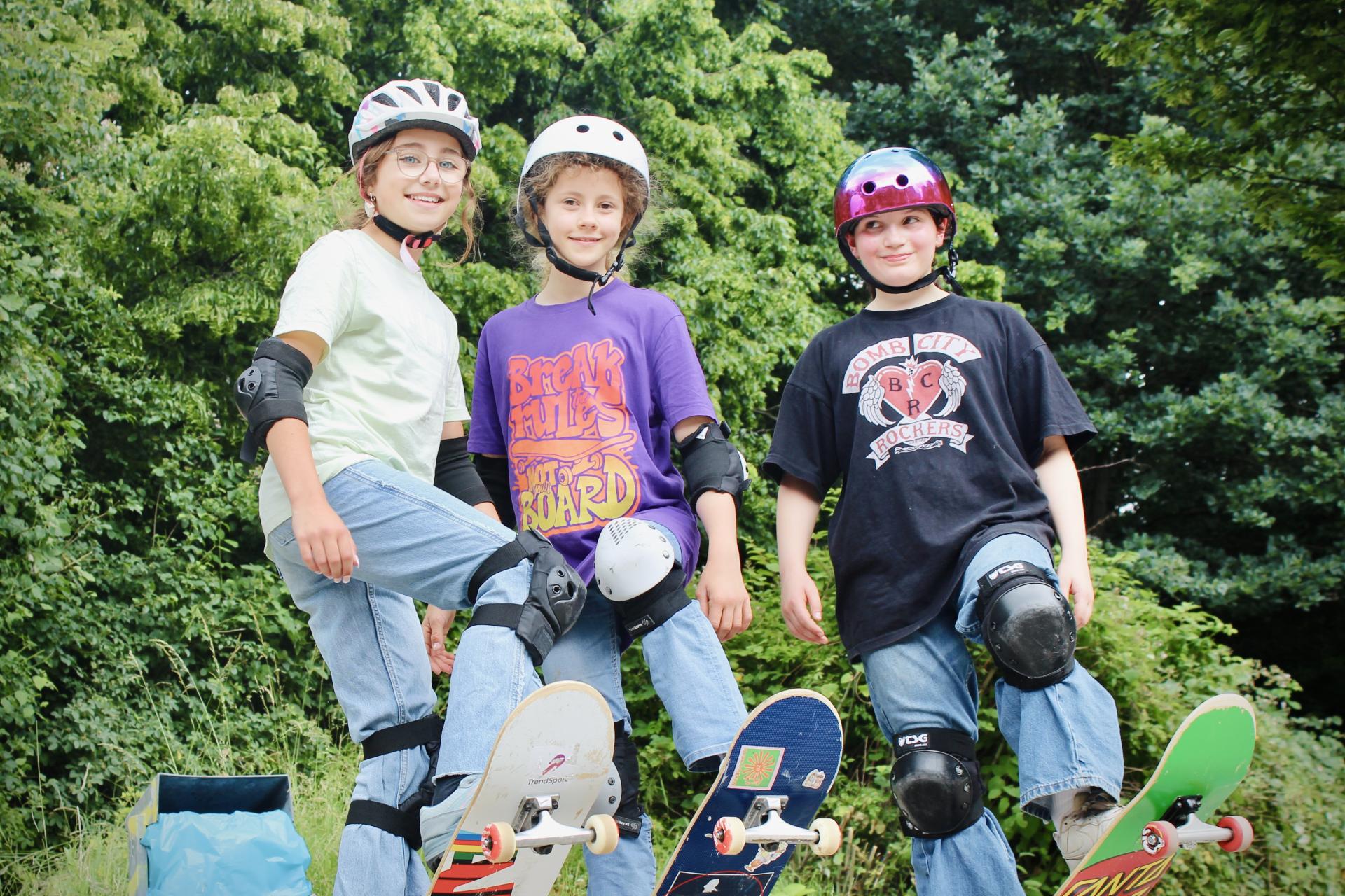 Drei Mädchen mit Helm und Schutzausrüstung stehen mit Skateboards im Grünen und posieren gemeinsam für ein Foto.