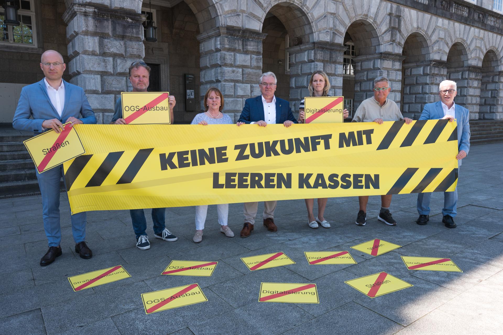 Oberbürgermeister, Kämmerer, Dezernent*innen, Bürgermeister*innen und Bezirksbürgermeister*innen halten ein gelb-schwarzes Banner mit der Aufschrift "Keine Zukunft mit leeren Kassen". Vor Ihnen liegen gelbe Schilder, auf denen Begriffe wie "Straßensanierung", "Digitalisierung" und "OGS-Ausbau" mit einem roten Balken durchgestrichen sind.