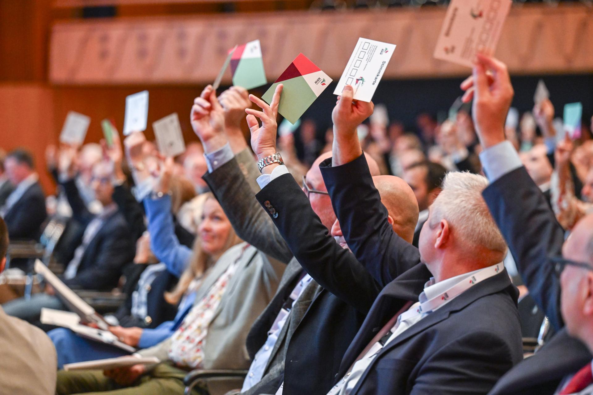Mehrere Personen sitzen in einem Saal und halten für eine Abstimmung Stimmzettel hoch.