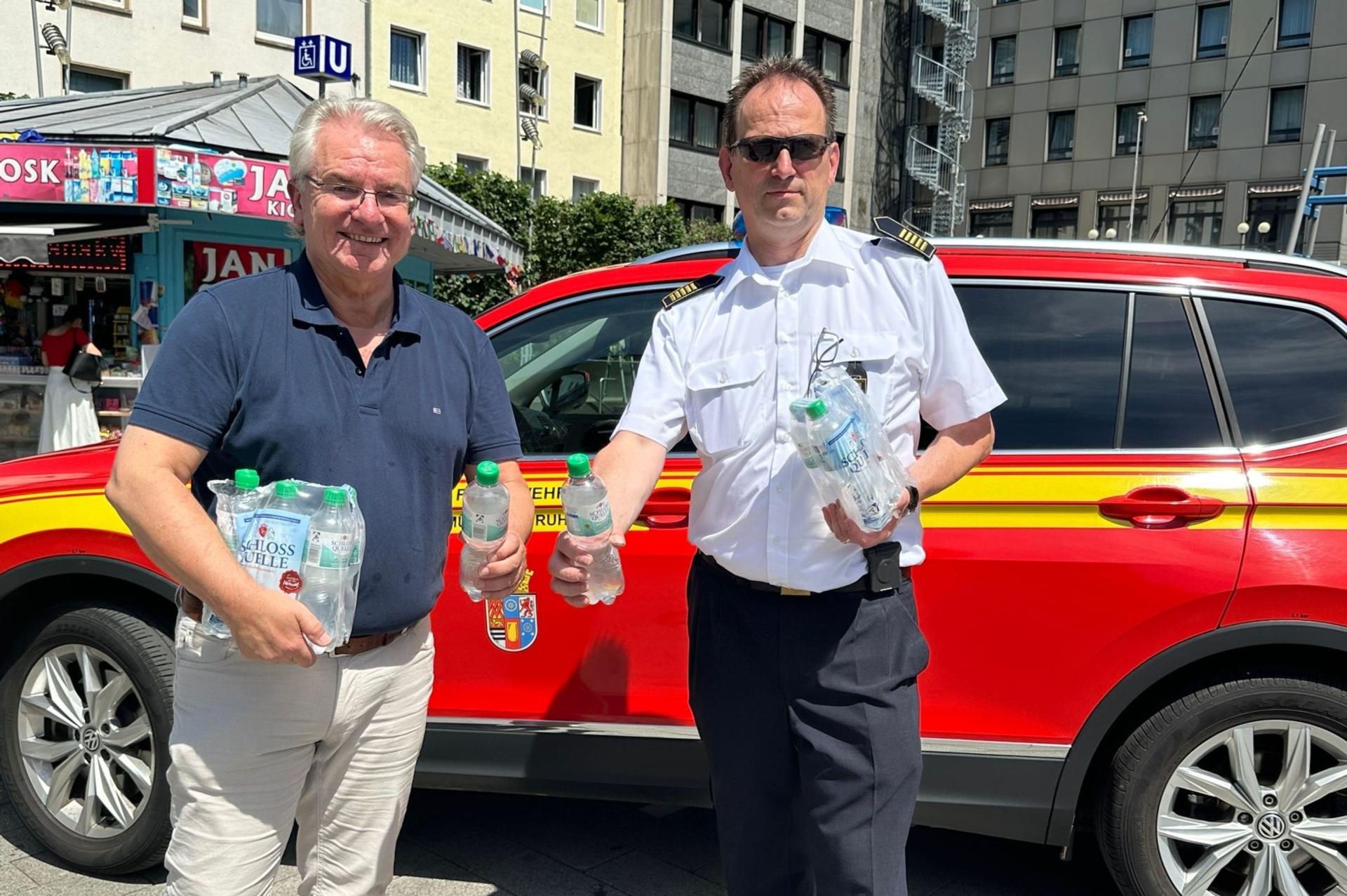 Der Oberbürgermeister und der Feuerwehrchef halten Träger mit Wasser im Arm und auch einzelne Wasserflaschen in der Hand. Sie stehen vor einem roten Feuerwehrauto. 