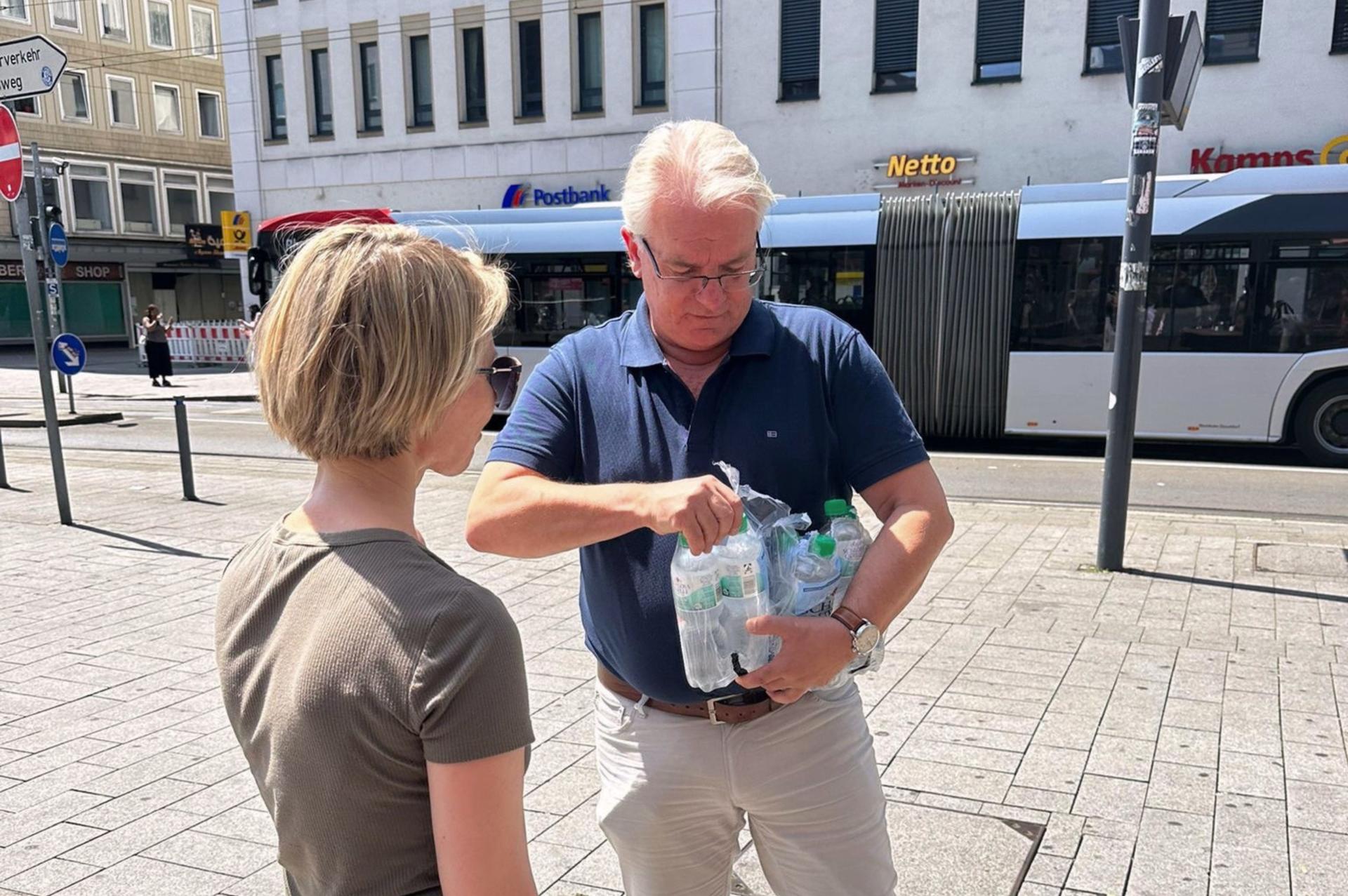 Der Oberbürgermeister hat einen Träger Wasser auf dem Arm und entnimmt eine Flasche, um sie einer vorbeikommenden Passantin zu überreichen.