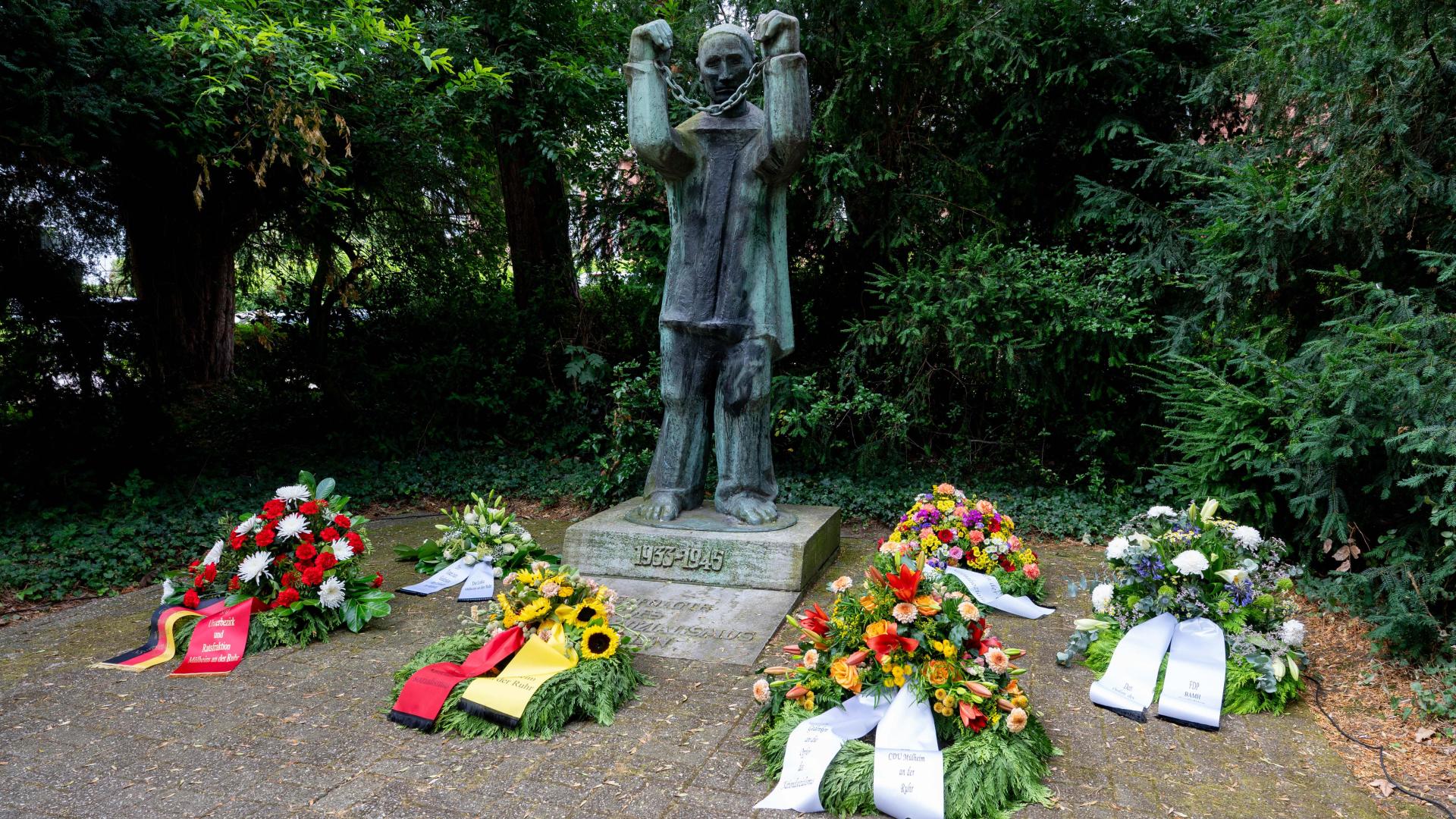 Im Hintergrund das Mahnmal. Es ist ein Mann, der seine mit einer Kette gefesselten Hände vor sich hoch hält. Um das Mahnmal sind Blumenkränze mit Schleifen abgelegt.