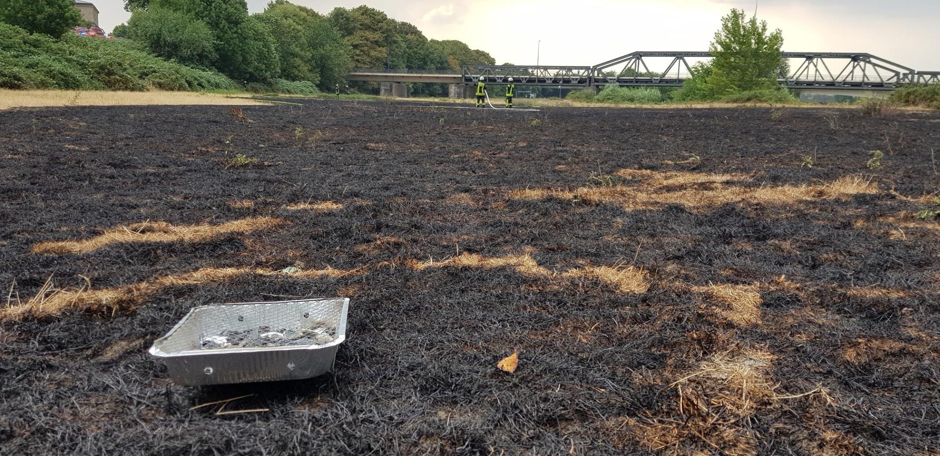 Im Vordergrund steht die Aluschale eines Wegwerfgrills auf einer verbrannten Wiese. Im Hintergrund sind zwei Feuerwehrmänner und die Raffelbergbrücke erkennbar.