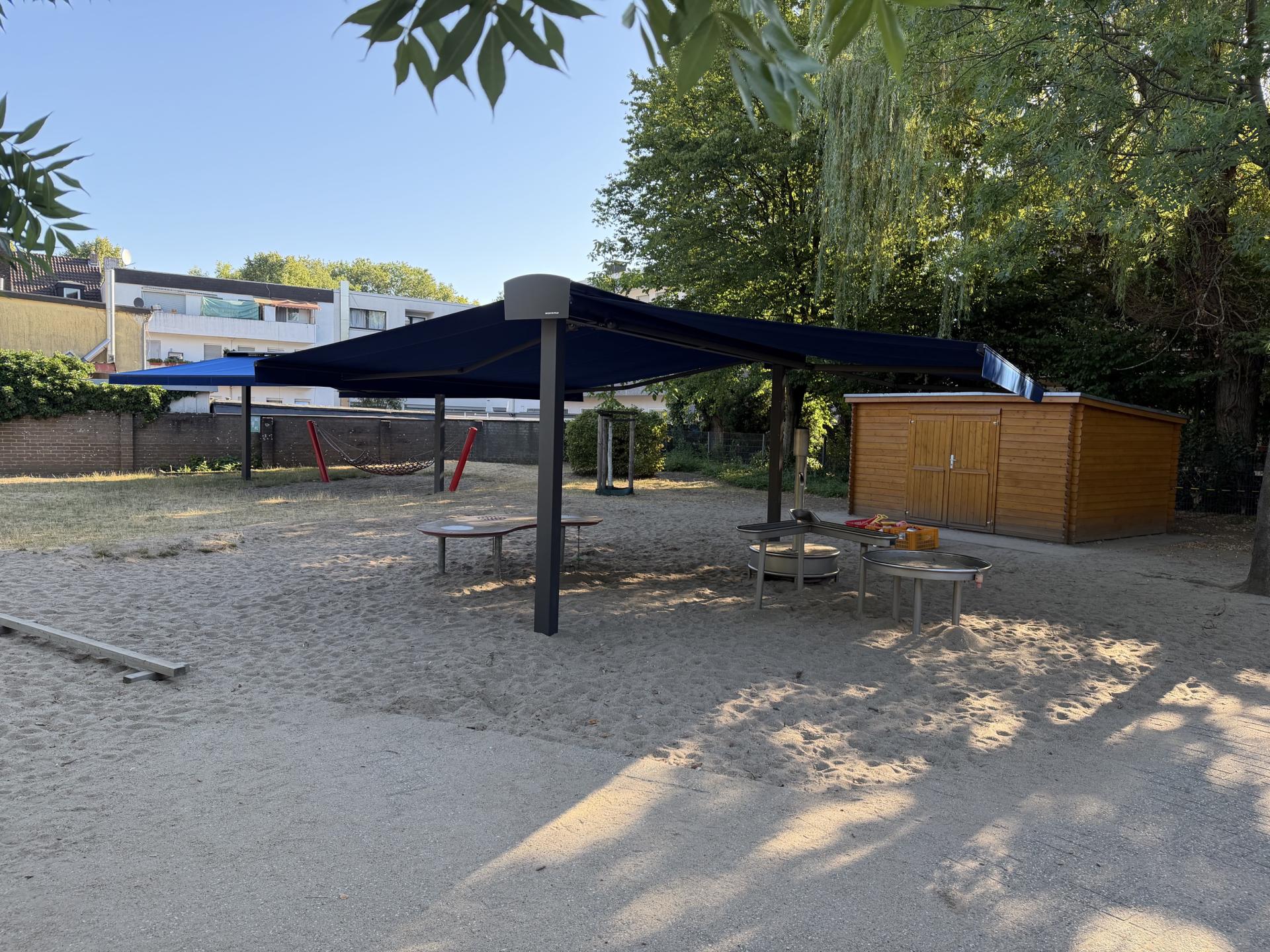 Im Außenbereich einer Kita sorgen zwei fest installierte Sonnensegel für ausreichend Schatten auf einer Sandfläche und einer Wiese. Im Hintergrund ein Blockhaus, Bäume und Wohnhäuser.