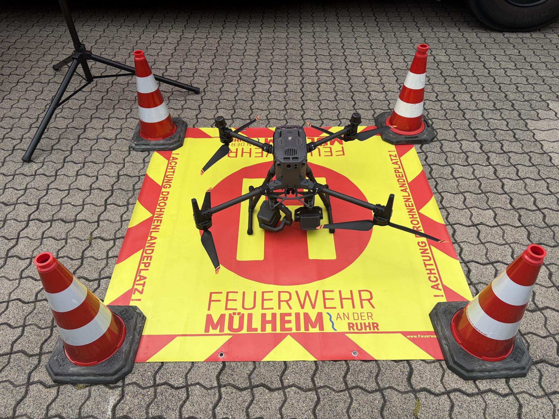 Auf einer gelb-roten quadratischen Bodenmarkierung, an vier Ecken mit Pylonen abgegrenzt, steht eine große Drohne.