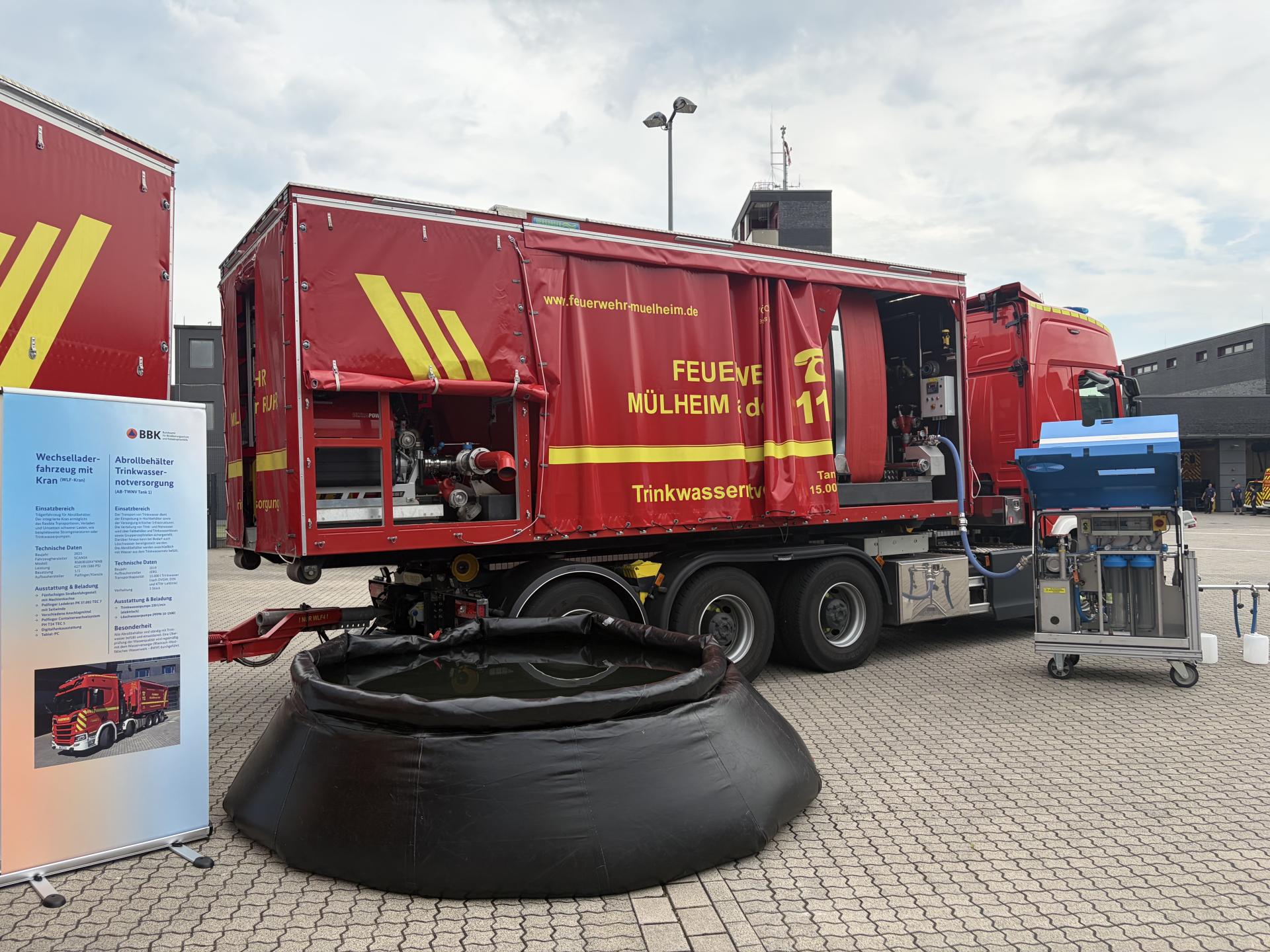 Ein LKW der Feuerwehr mit Anhänger zur Trinkwassernotversorgung. Davor ist ein Roll-up mit Informationen aufgebaut. Außerdem ein schwarzes aufblasbares Becken gefüllt mit Wasser.
