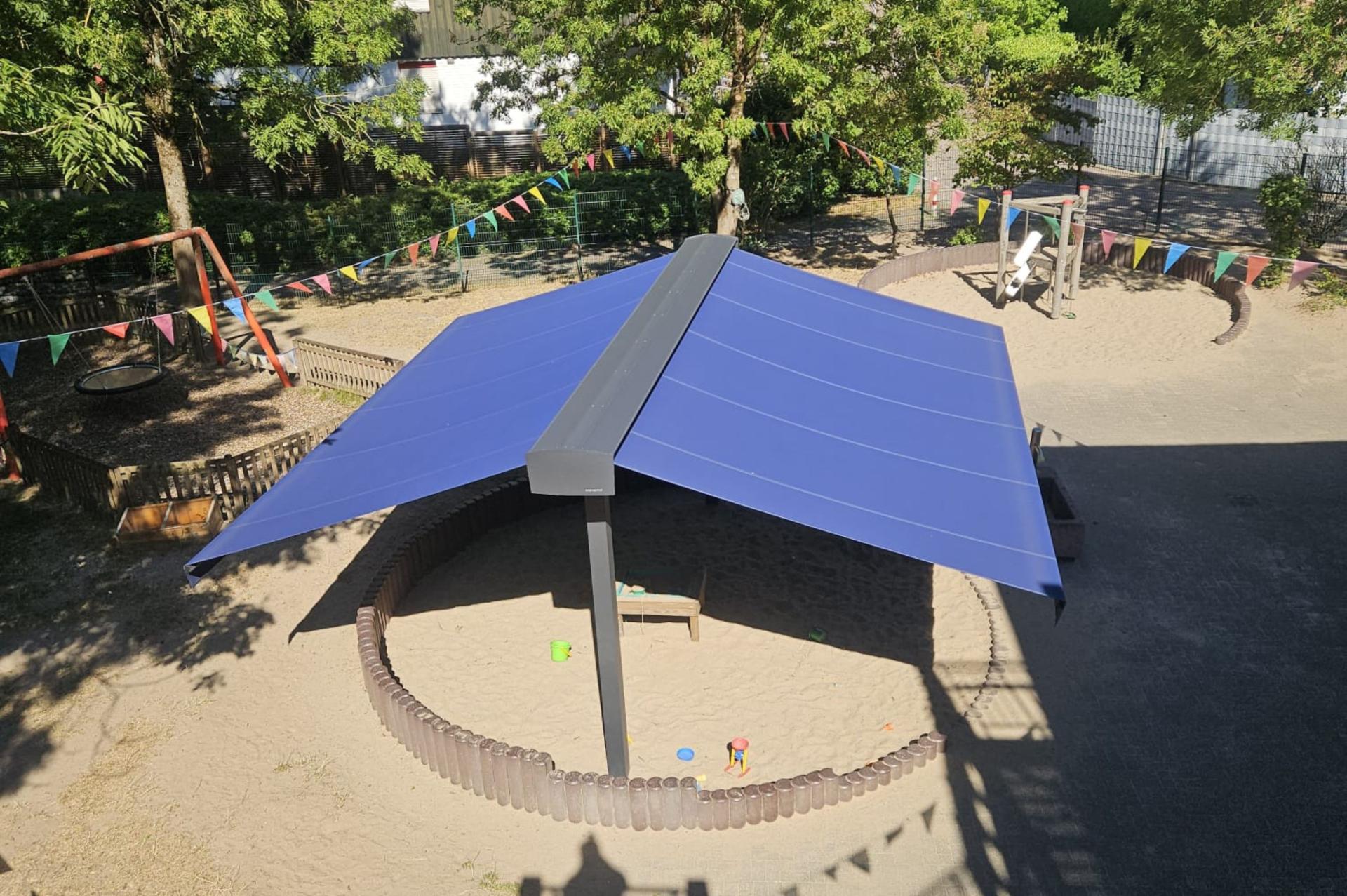 Die runde Sandspielfläche einer Kita wird von einem festinstallierten Sonnenschutz überspannt. Von einer Mittelstrebe aus spannen sich zwei blaue Sonnensegel nach links und nach rechts.