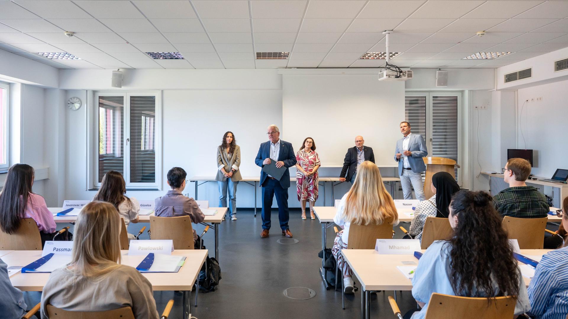 Mehrere Personen sitzen in der Feuerwehrschule und hören vier stehenden Personen zu, die vorne sprechen.