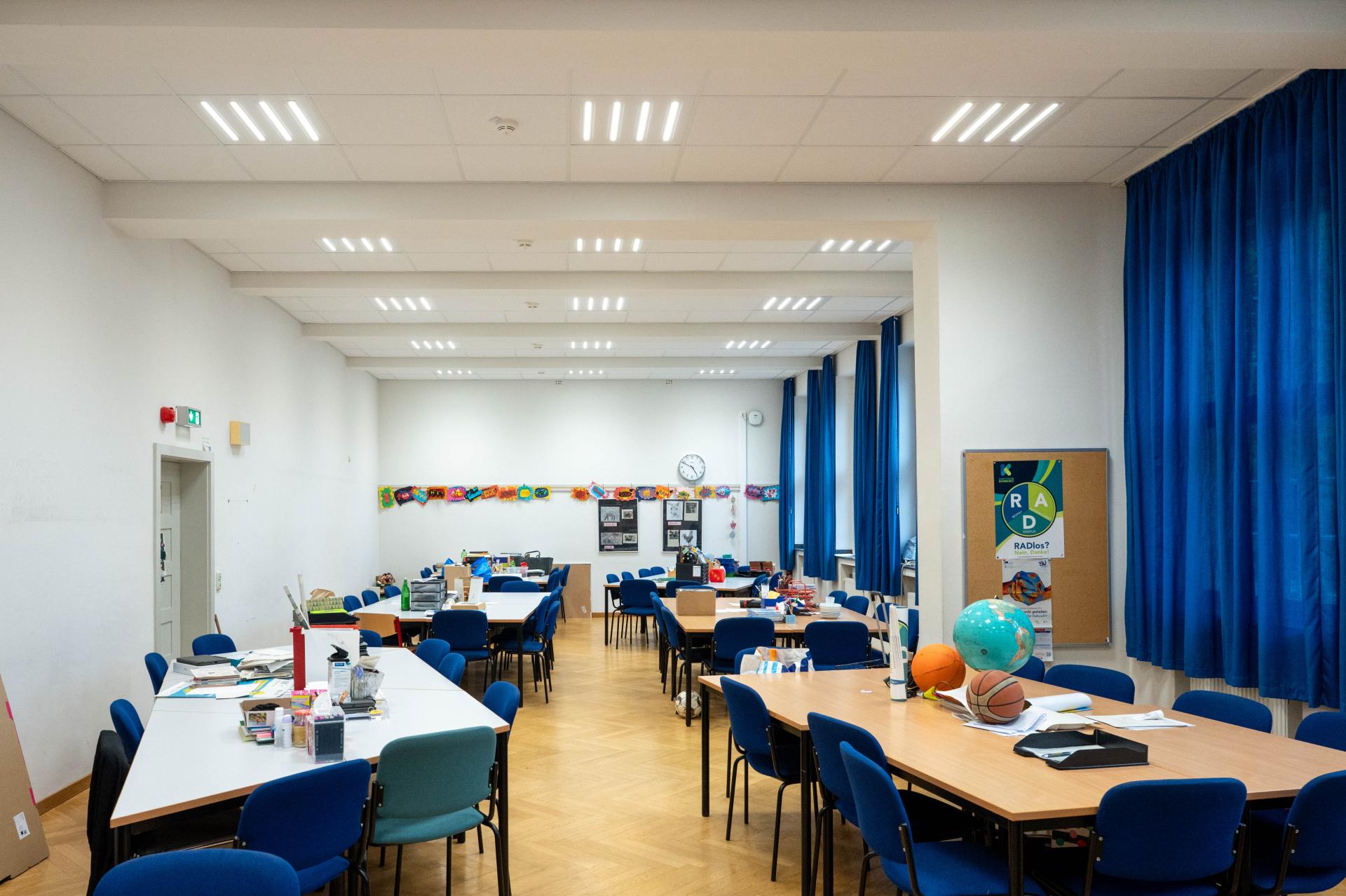 Das Bild zeigt ein Lehrerzimmer mit vielen Tischen und Stühlen. An den Wänden hängen bunte Kinderzeichnungen und eine Pinnwand mit einem Poster. Blaue Vorhänge bedecken die Fenster.