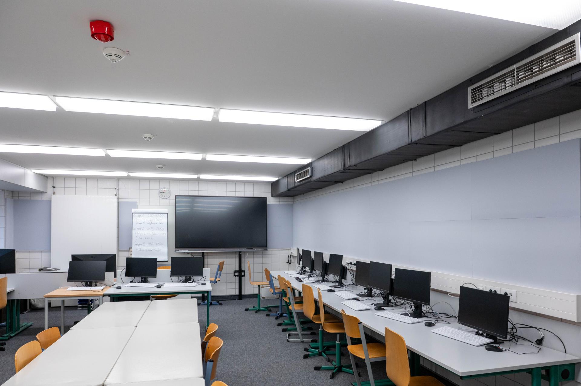 Dies ist ein Computerraum mit Reihen von Computern und Sitzplätzen. An der Wand hängt ein grosser Bildschirm.