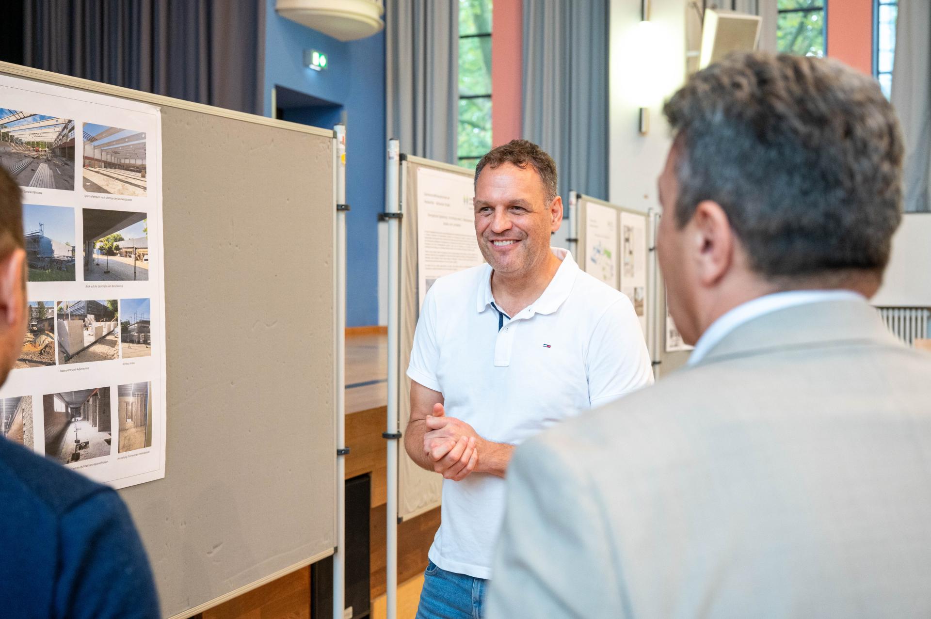 Ein Mann in einem weißen Poloshirt steht vor einer Pinnwand mit Bildern und spricht mit einer anderen Person. Im Hintergrund ist eine weitere Person zu sehen, die die Präsentation betrachtet.