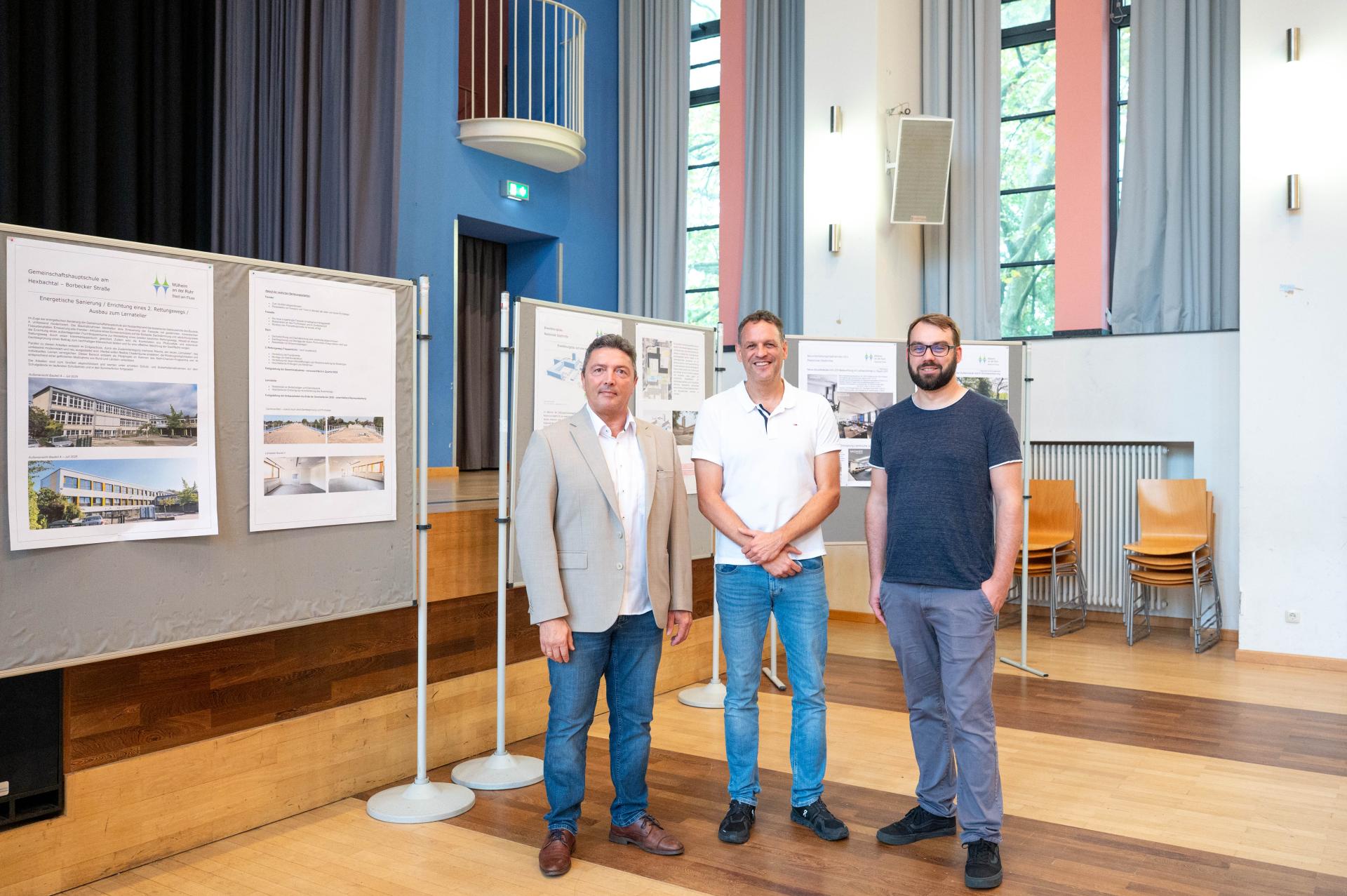 Drei Männer stehen vor mehreren Ausstellungstafeln, die Informationen und Bilder über ein Schulprojekt zeigen.