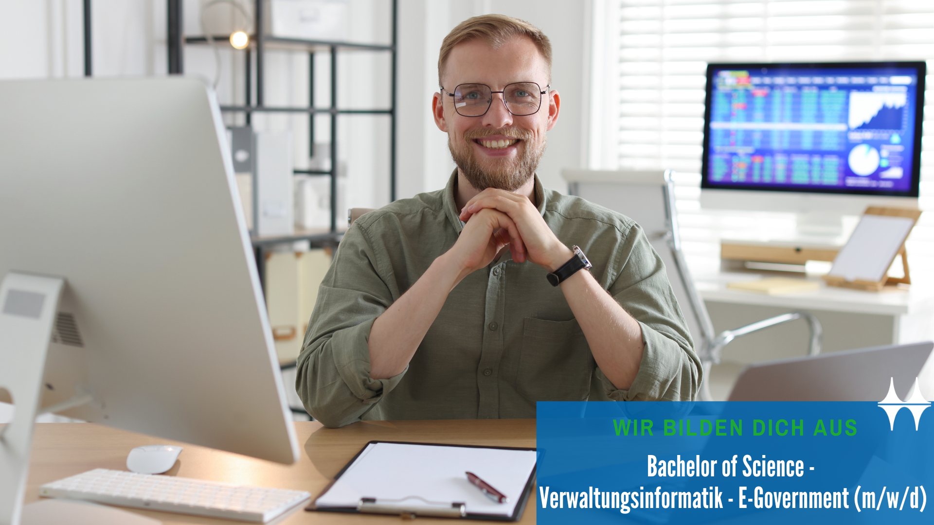 Auf dem Bild sitzt ein Mann an einem Schreibtisch. Vor ihm links steht ein Bildschirm. Der Mann lächelt in die Kamera.