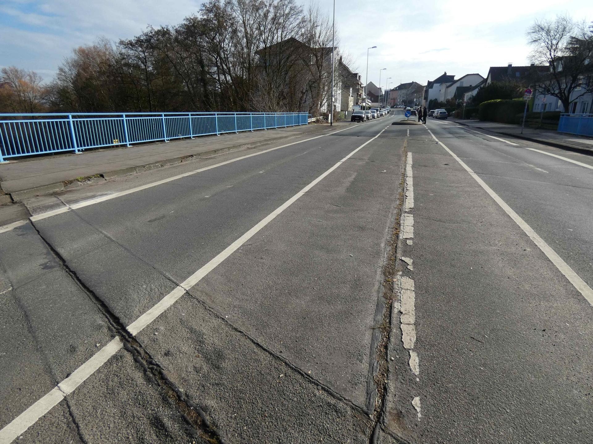 Dargestellt sind Fahrbahn und Gehwege, die über die Brücke führen. Links ist ein blaues Brückengeländer erkennbar. Der Asphalt ist sichtbar geschädigt. Im Straßenverlauf sind parkende Autos sowie links und rechts der Straße Häuser und Bäume abgebildet.
