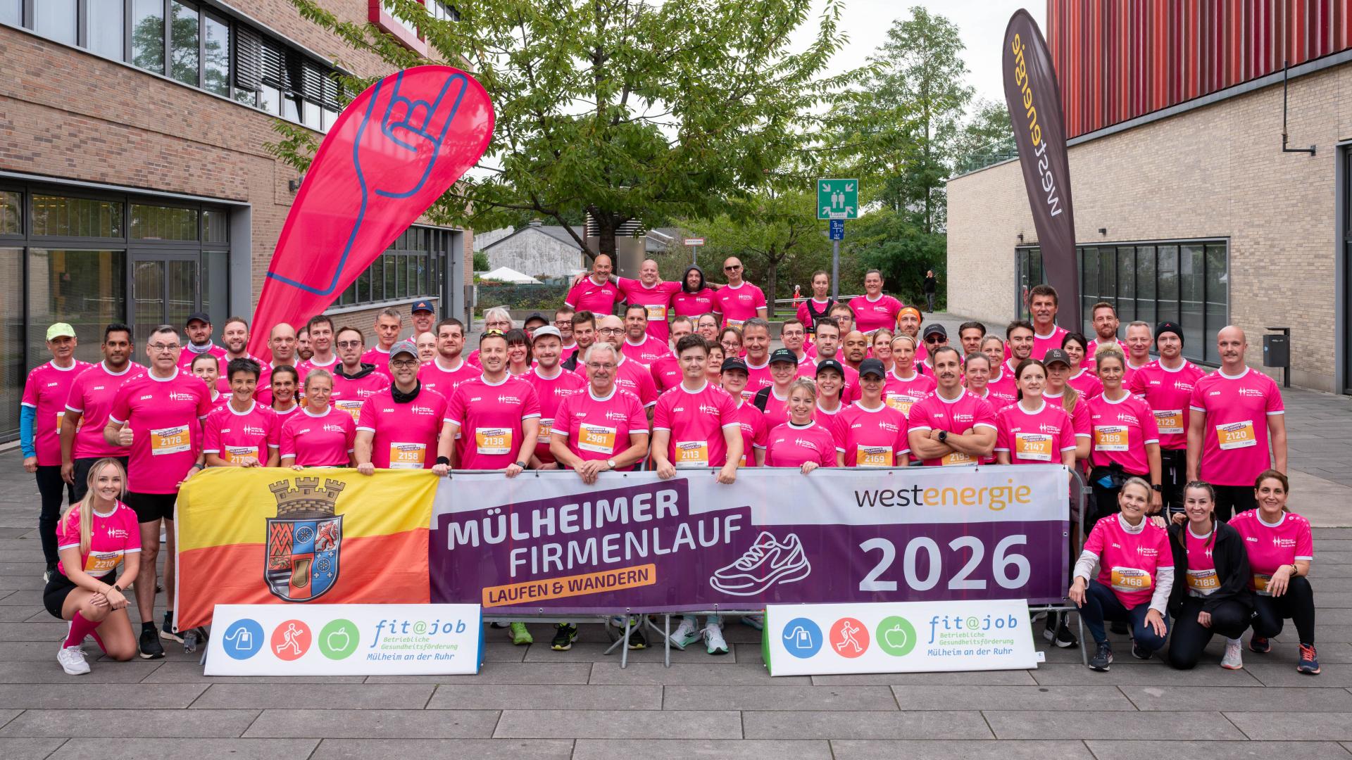 Große Gruppe von Menschen in pinken Laufshirts steht vor Gebäuden und hält ein Banner mit der Aufschrift 'MÜLHEIMER FIRMENLAUF 2026'