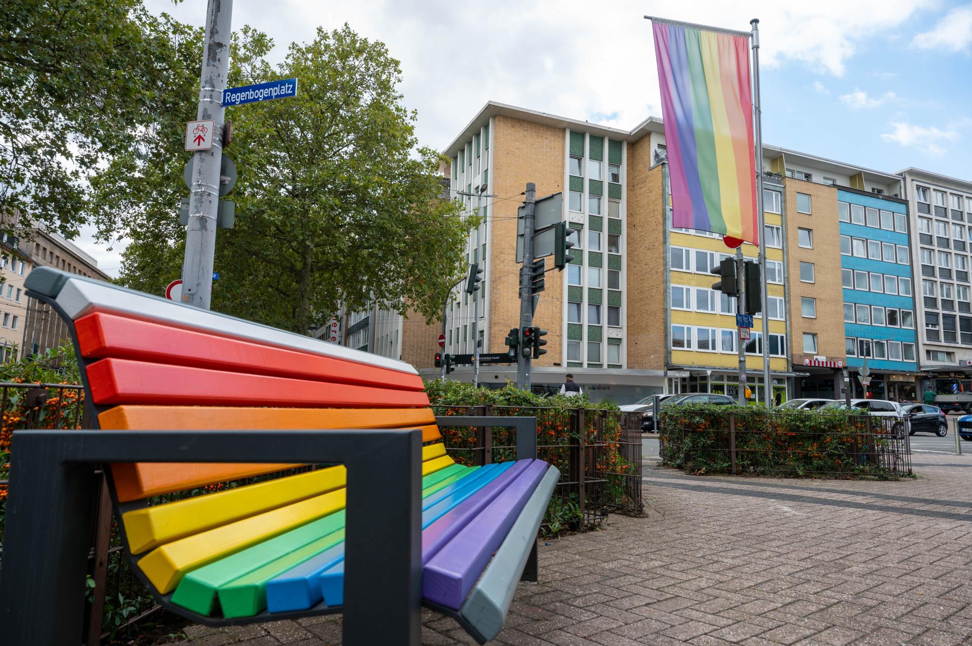Im Vordergrund eine regenbogenfarbene Sitzbank. Dahinter ein Laternenmast mit dem Straßenschild "Regenbogenplatz". Es weht an einem Mast außerdem eine Regenbogenflagge. Im Hintergrund Häuser und Bäume.