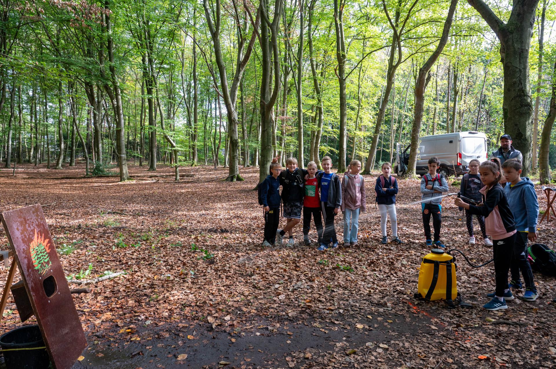 Eine Schulkasse steht auf einer Lichtung in einem alten Buchenwald. Die Klasse feuert eine Mitschülerin an, die mit einer Wasserspritze in einen Eimer hinter einem Schaubild mit einem Waldbrand zielt.