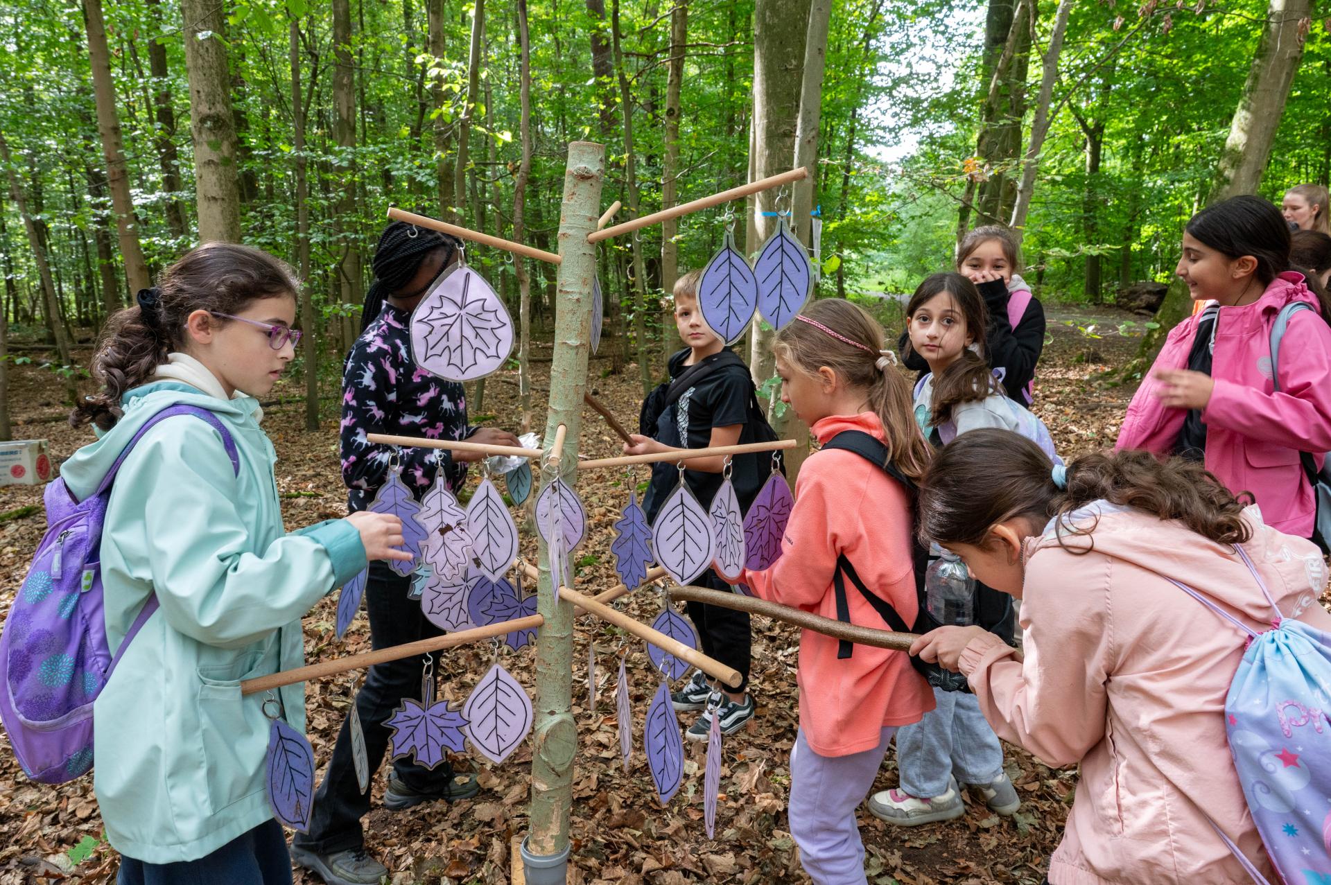 An ein Holzgestell mit Haken werden von einer Gruppe von Kindern Blätter verschiedene Blätter angebracht.
