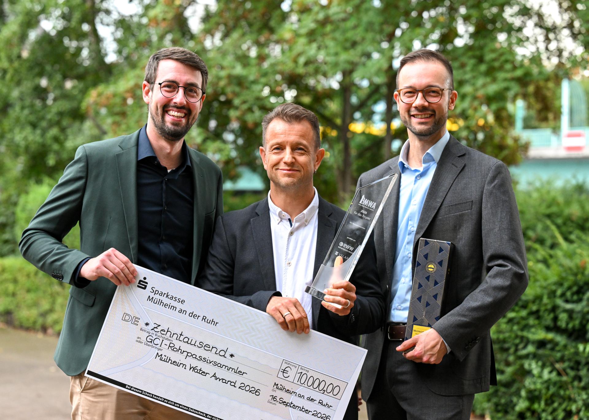Die drei Preisträger des Mülheimer Water Awards halten den Preis und einen Scheck in der Hand. Sie stehen an der Ruhrpromenade vor der Stadthalle in Mülheim.