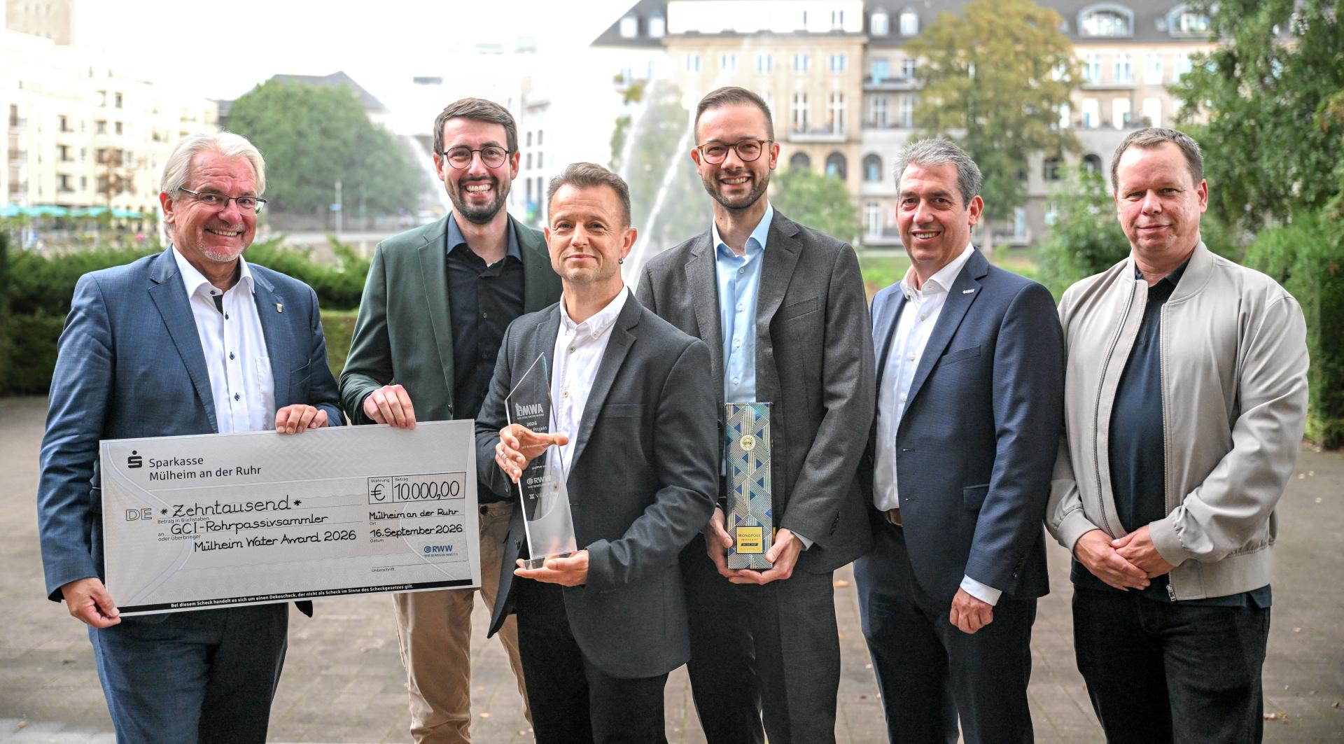 Das Foto zeigt den Oberbürgermeister der Stadt Mülheim, Marc Buchholz, bei der Verleihung des Mülheimer Water Awards mit den Siegern vor der Stadthalle. Im Hintergrund ist die Ruhrpromenade zu sehen. Die Preisträger halten den Pokal und einen Scheck in der Hand.