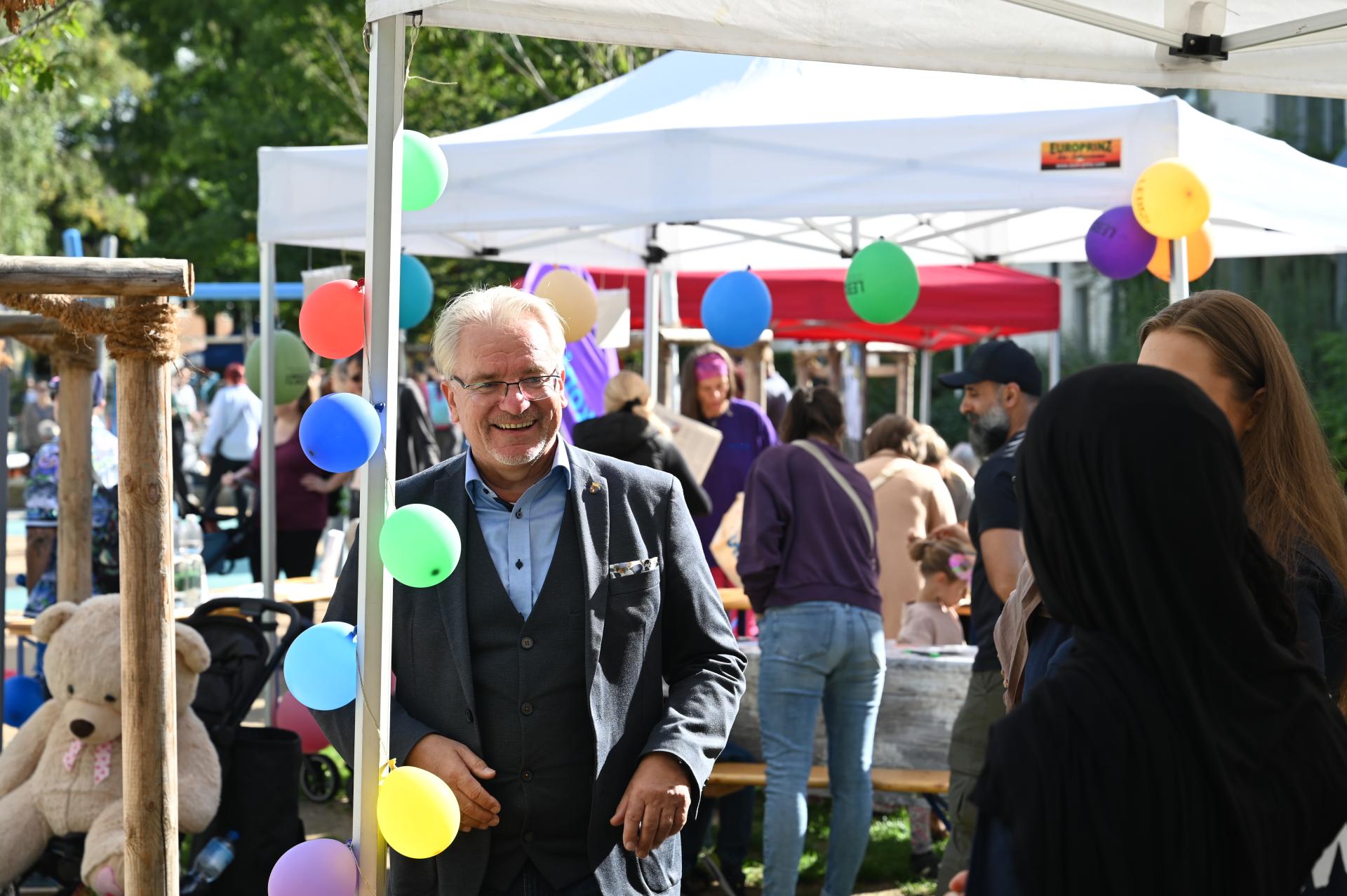 Der Oberbürgermeister tritt an einen mit Luftballons geschmückten Pavillon. Im Hintergrund sind weitere Stände auf dem Spielplatzfest am Schloßberg zu erkennen.
