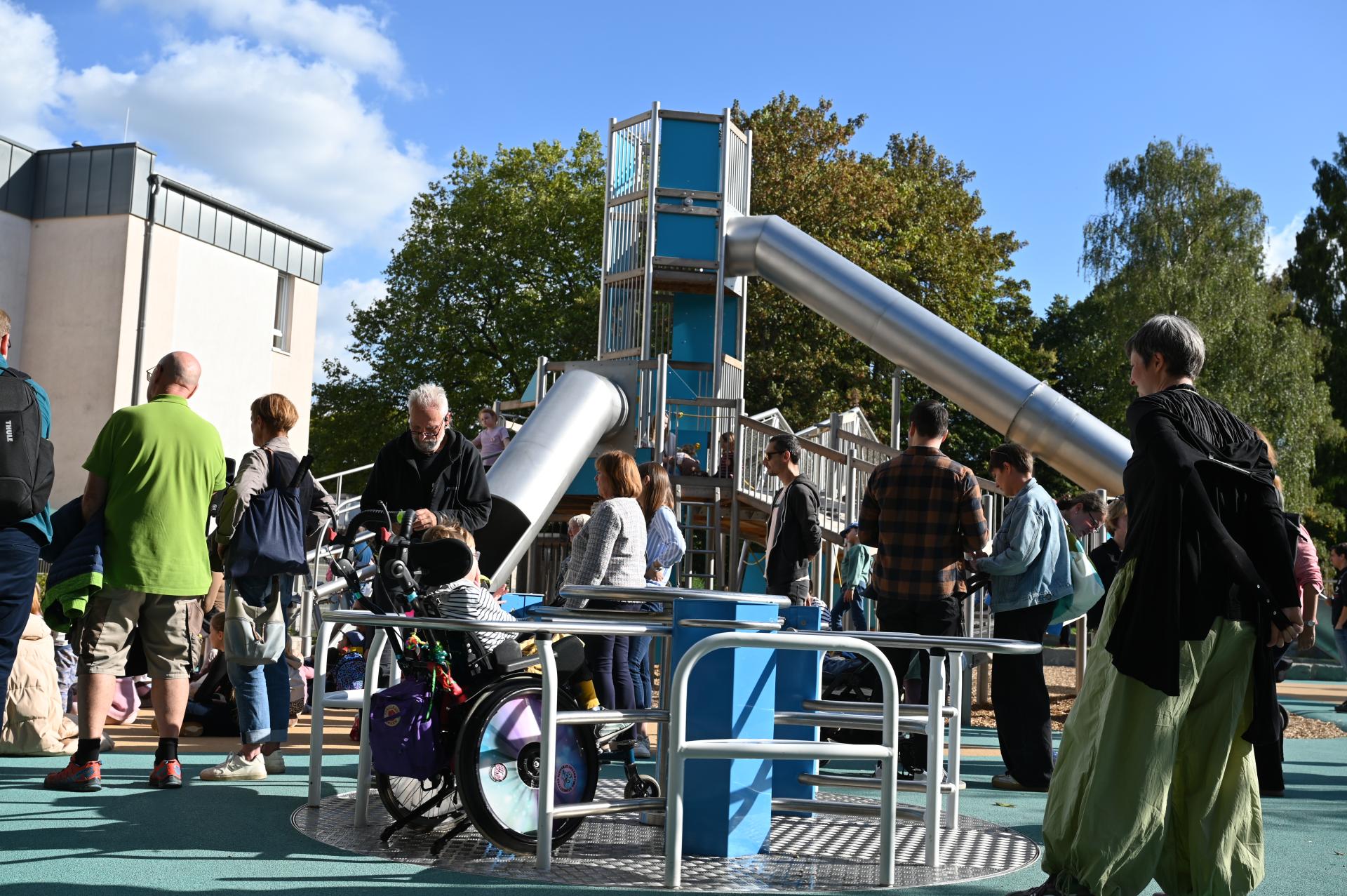 Im Vordergrund das Karussell auf dem Inklusiven Spielplatz am Schloßberg, das beim Spielplatzfest gerade von einem Kind mit Rollstuhl genutzt wird. Im Hintergrund das Großspielgerät und die vielen Gäste der Einweihungsfeier.