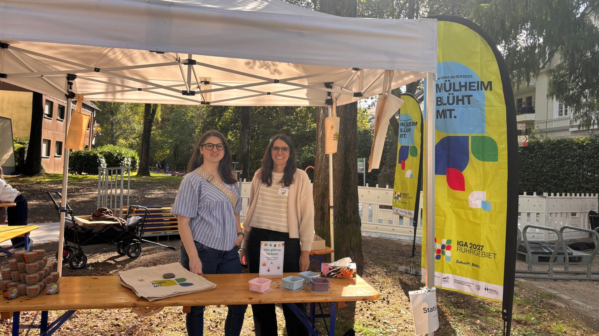 Das Foto zeigt den Stand des Quartiersbüros bei der Eröffnungsfeier des Spielplatzes am Schloßberg, es sind zwei Frauen hinter einem Tisch zu sehen mit einer Beachflag zur IGA in Mülheim