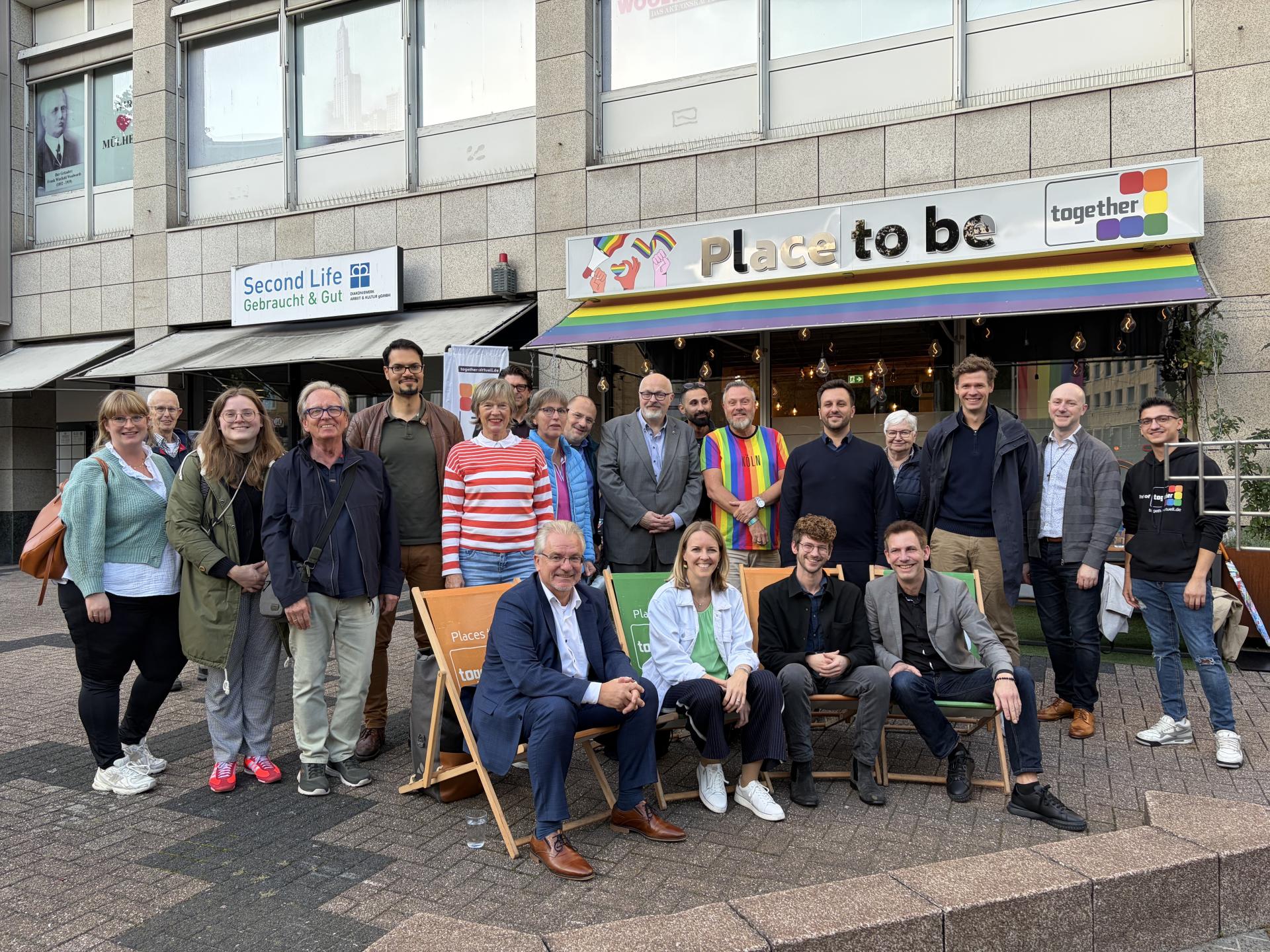 Gruppenfoto vor dem SVLS e.V. Ladenlokal am neuen Regenbogenplatz. In der vorderen Reihe sitzen vier Personen auf Liegestühlen - ganz links Oberbürgermeister Marc Buchholz. Weitere Personen stehen dahinter.