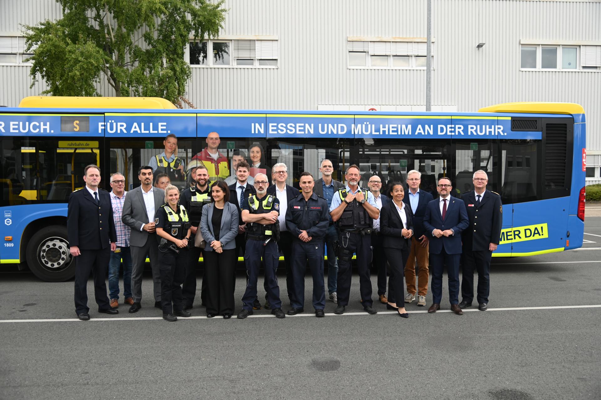 Gruppe von Menschen, darunter uniformierte Sicherheitskräfte und in Anzügen gekleidete Personen, steht vor einem blauen Bus mit gelbem Schriftzug