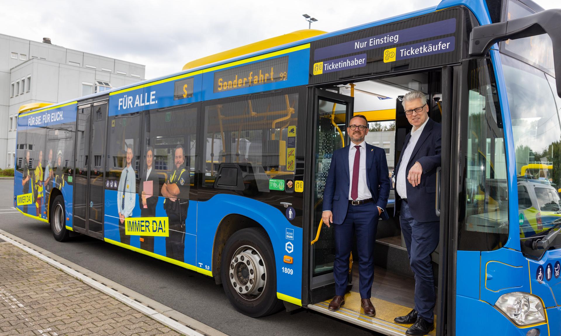 Zwei Männer in Anzügen stehen in der geöffneten Tür eines blauen Stadtbusses mit Werbeaufschrift und mehreren abgebildeten Personen an der Seite