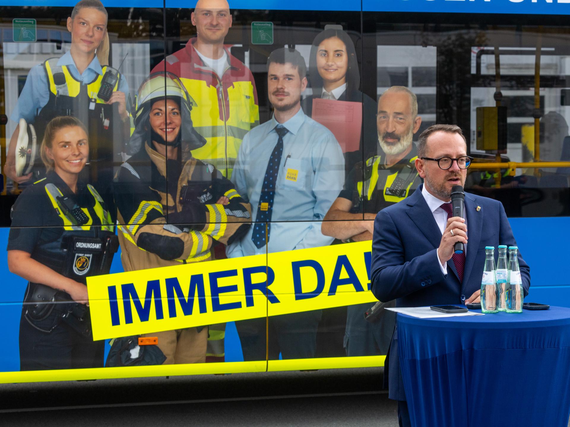 Mann mit Brille spricht in Mikrofon vor blauem Tisch mit Wasserflaschen, im Hintergrund großes Werbeplakat mit verschiedenen Berufsgruppen und gelbem Schriftzug 'IMMER DA'