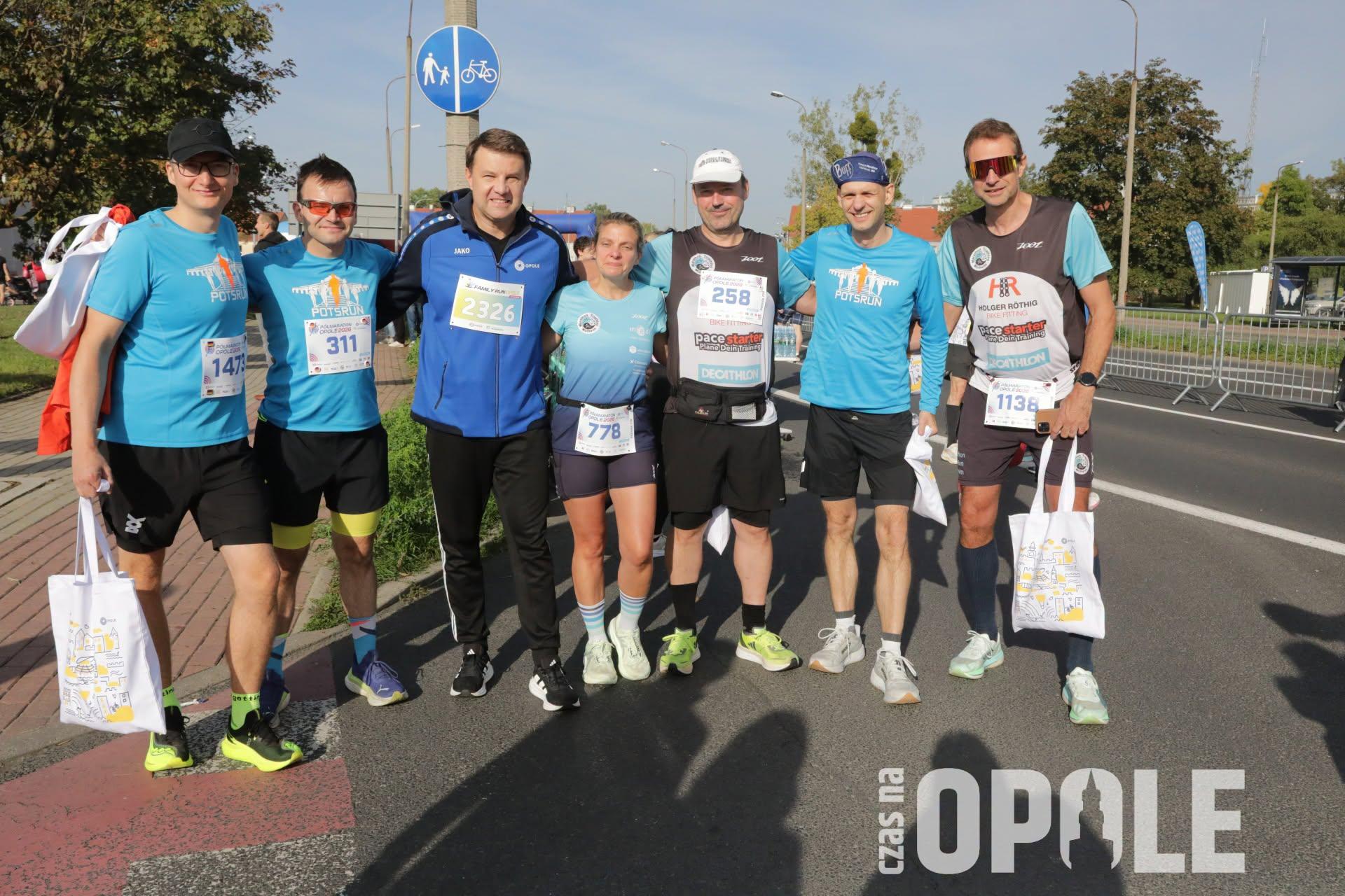 Sieben Personen in Sportkleidung haben sich für ein Foto aufgestellt. Sie halten sich in den Armen. Alle haben Startnummern auf der Kleidung. Sie stehen auf einer abgesperrten Straße. Im Hintergrund einige Häuser und etwas Grün.