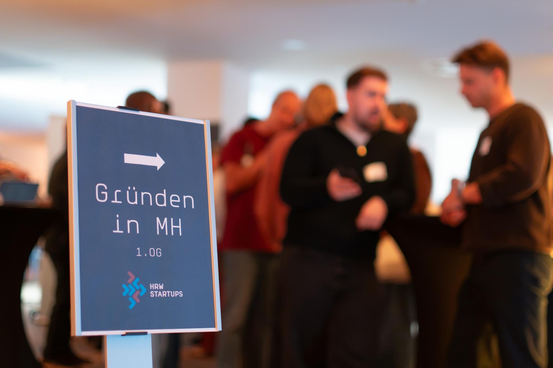 Auf dem Bild sieht man einen Aufsteller mit dem Hinweis "Gründen in Mülheim" am 14. Oktober 2026 und im Hintergrund eine kleine Menschenmenge