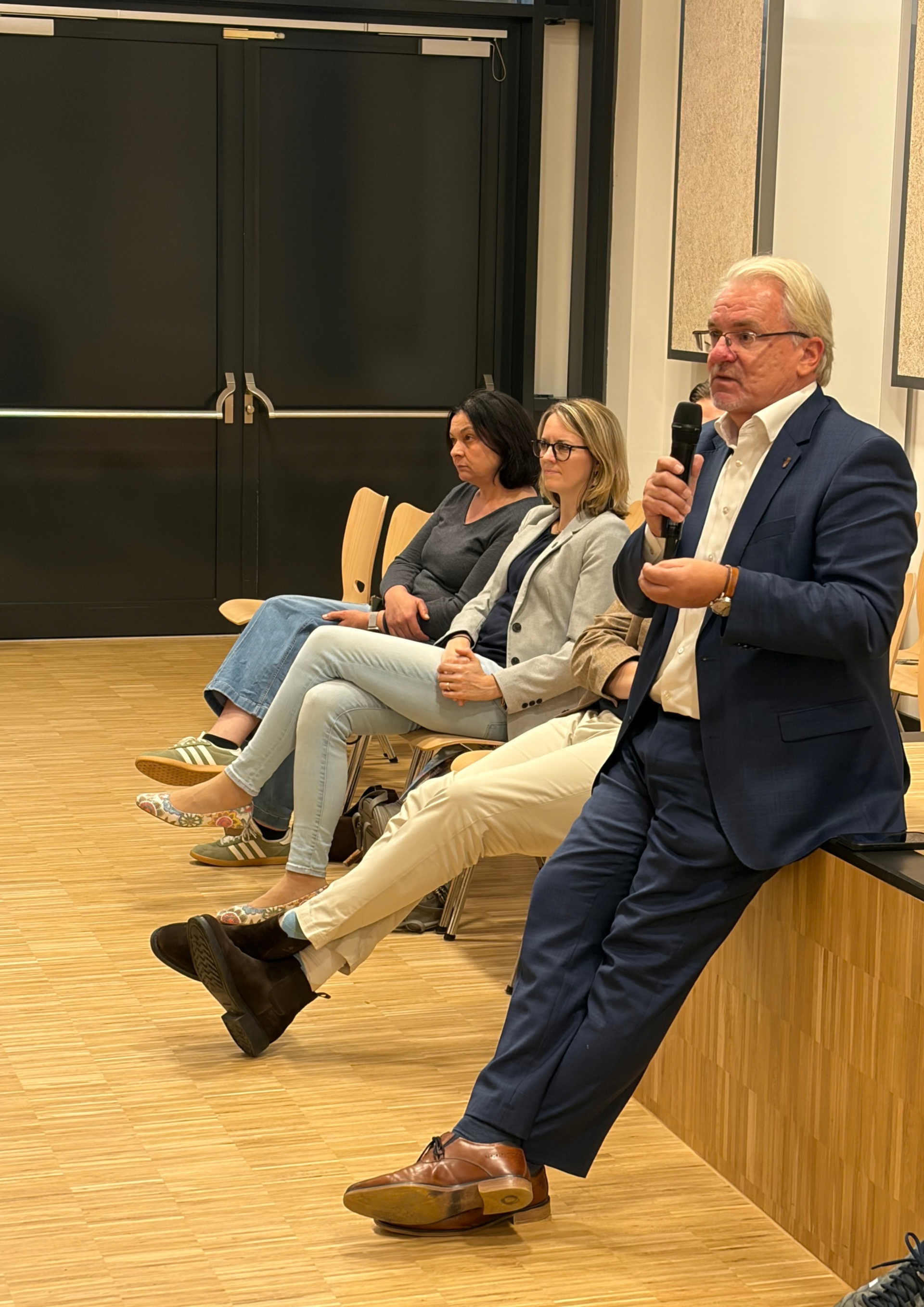 Oberbürgermeister mit Mikrophon lehnt auf einem Podest und spricht. Im Hintergrund mehrere Personen, die bei der Veranstaltung im Publikum sitzen.